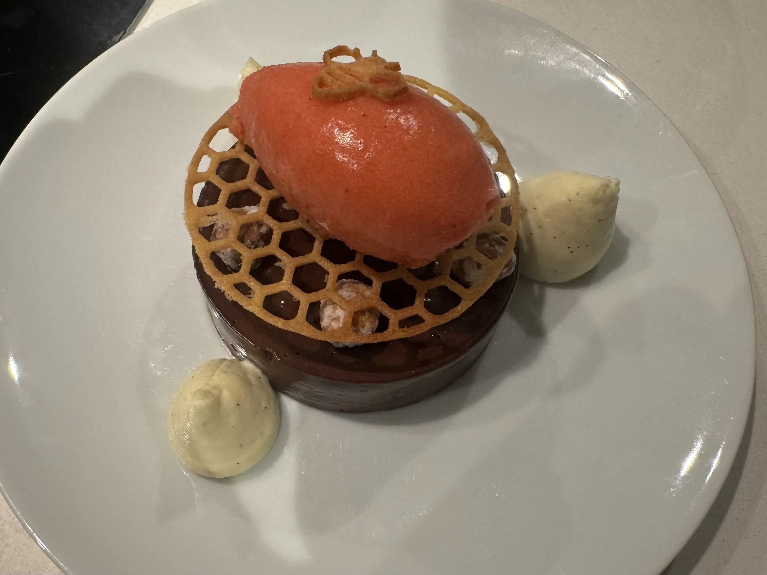A plated dessert featuring a round chocolate component topped with a honeycomb-patterned tuile, a scoop of red fruit gelée, two dollops of white cream, and small garnishes of caramel and piped cream.