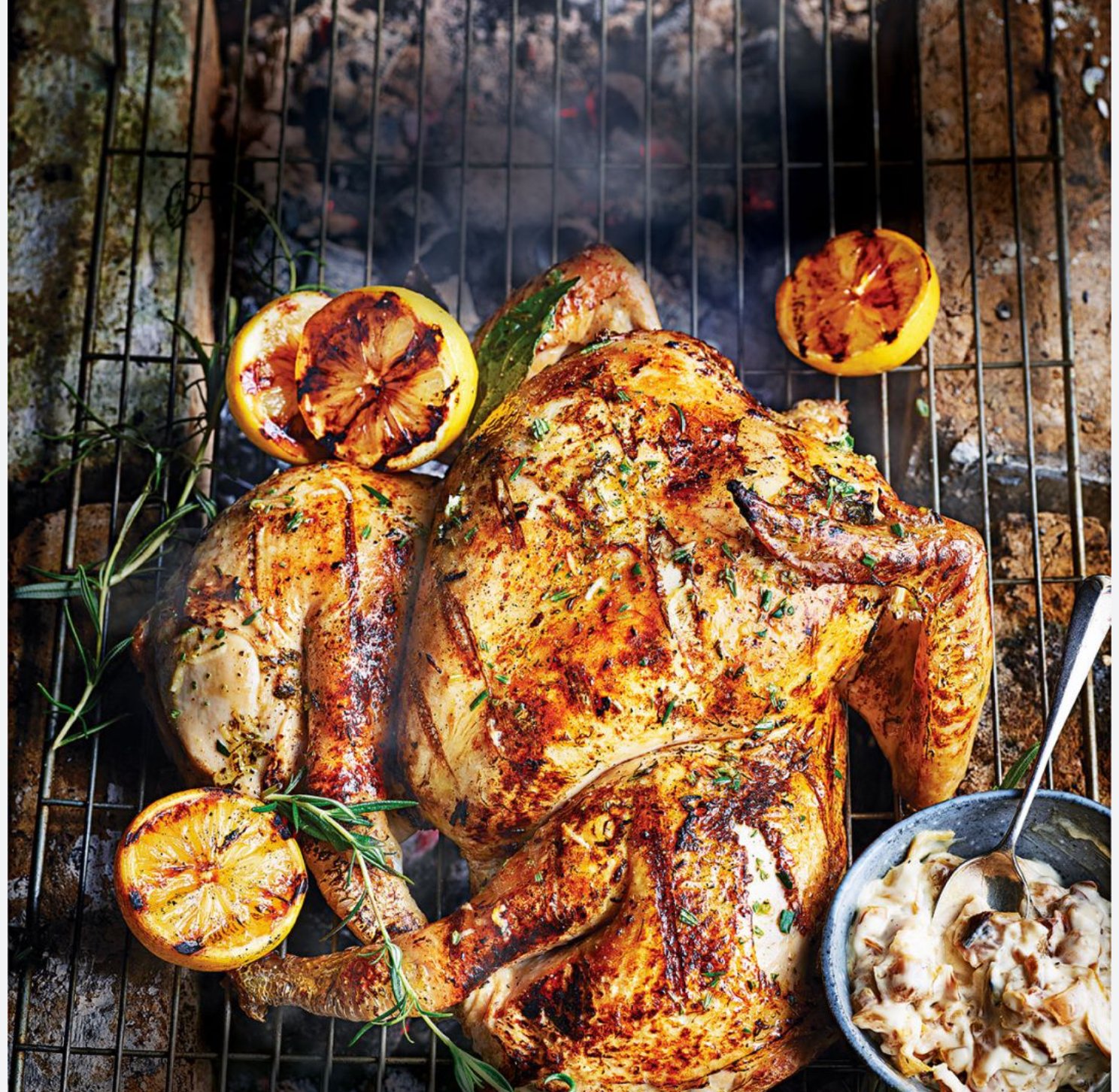 Grilled whole chicken with charred lemon halves and fresh herbs on a rack over a fire, with a side bowl of mushroom sauce.