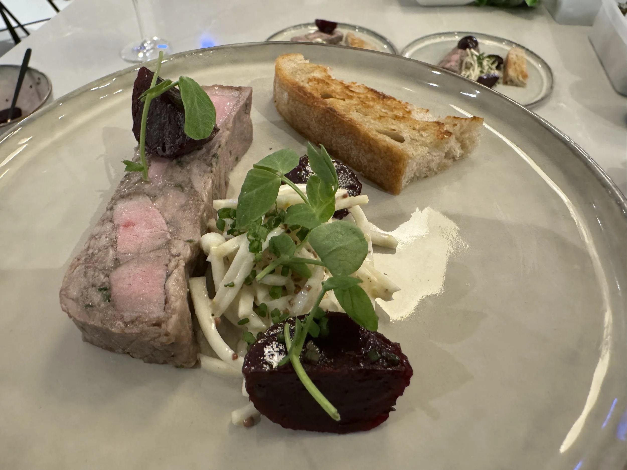 A plate with a slice of cooked meat, a piece of toasted bread, white pasta with green herbs, and red sauce or garnish, served on a white tablecloth.