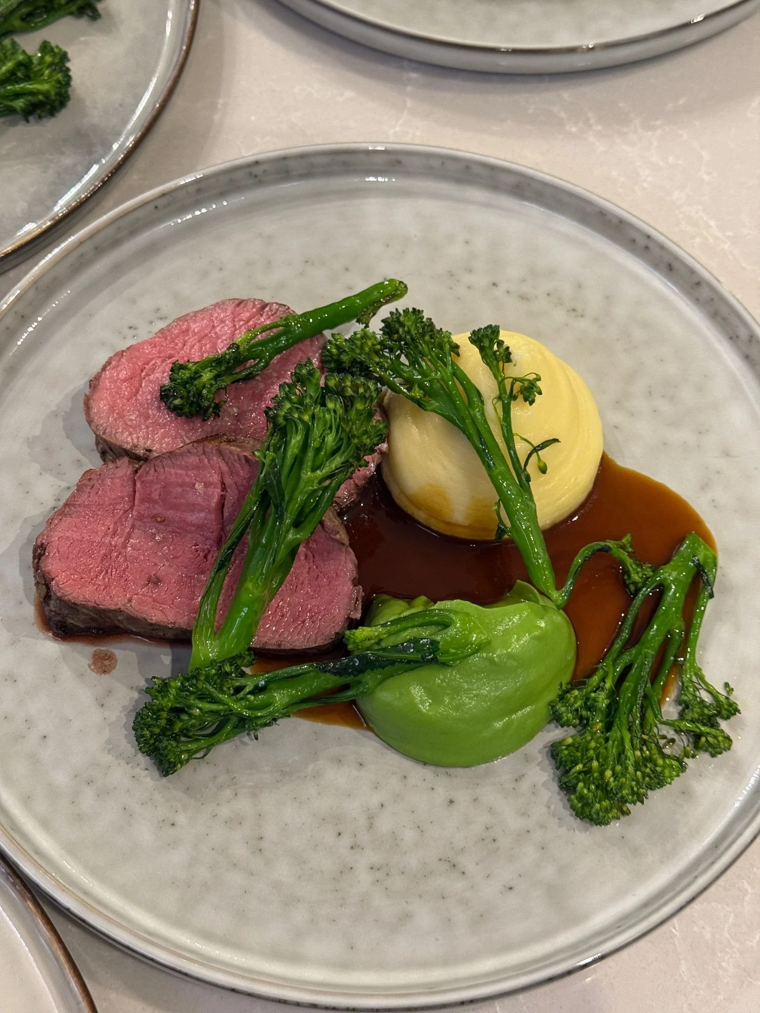 Slices of cooked beef, broccolini, a dollop of mashed potatoes, and sauce on a white plate.