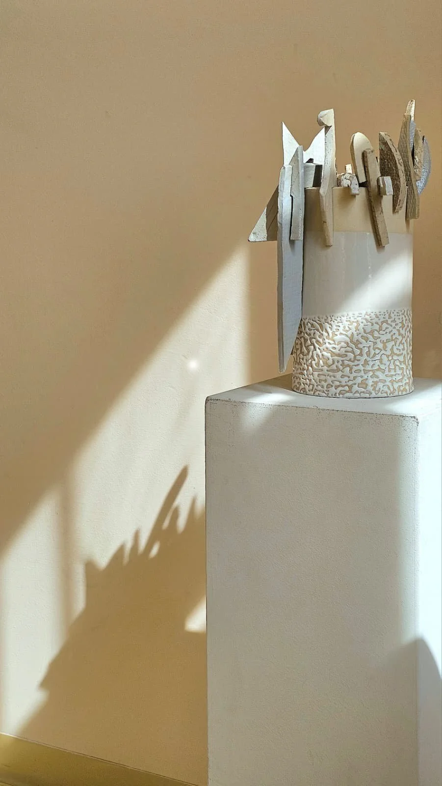 A white ceramic vase with a textured gold and white pattern holding wooden art pieces, placed on a white pedestal against a beige wall, with sunlight casting shadows.