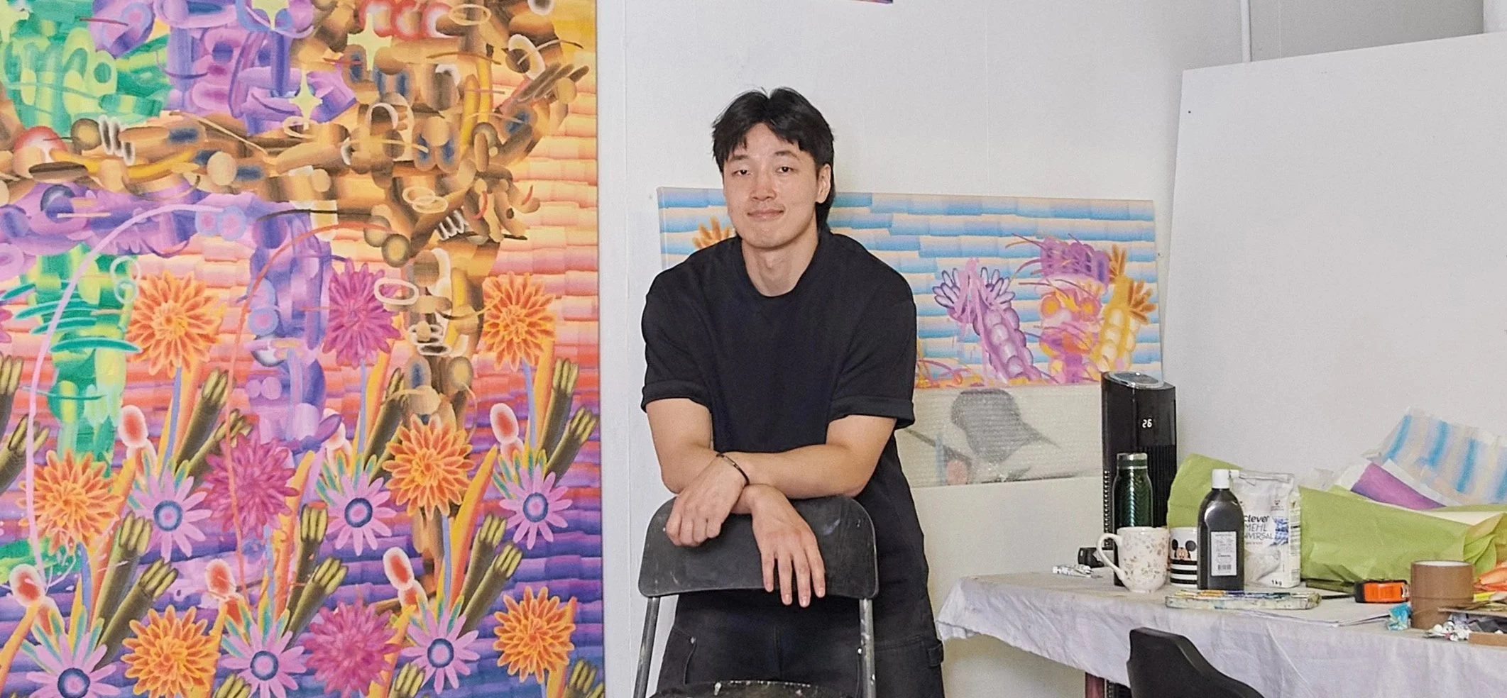 A young man in a black T-shirt stands indoors with his arms crossed on a black chair, next to colorful, abstract artwork of flowers and geometric patterns on a wall. There is a table with art supplies and a workspace behind him.