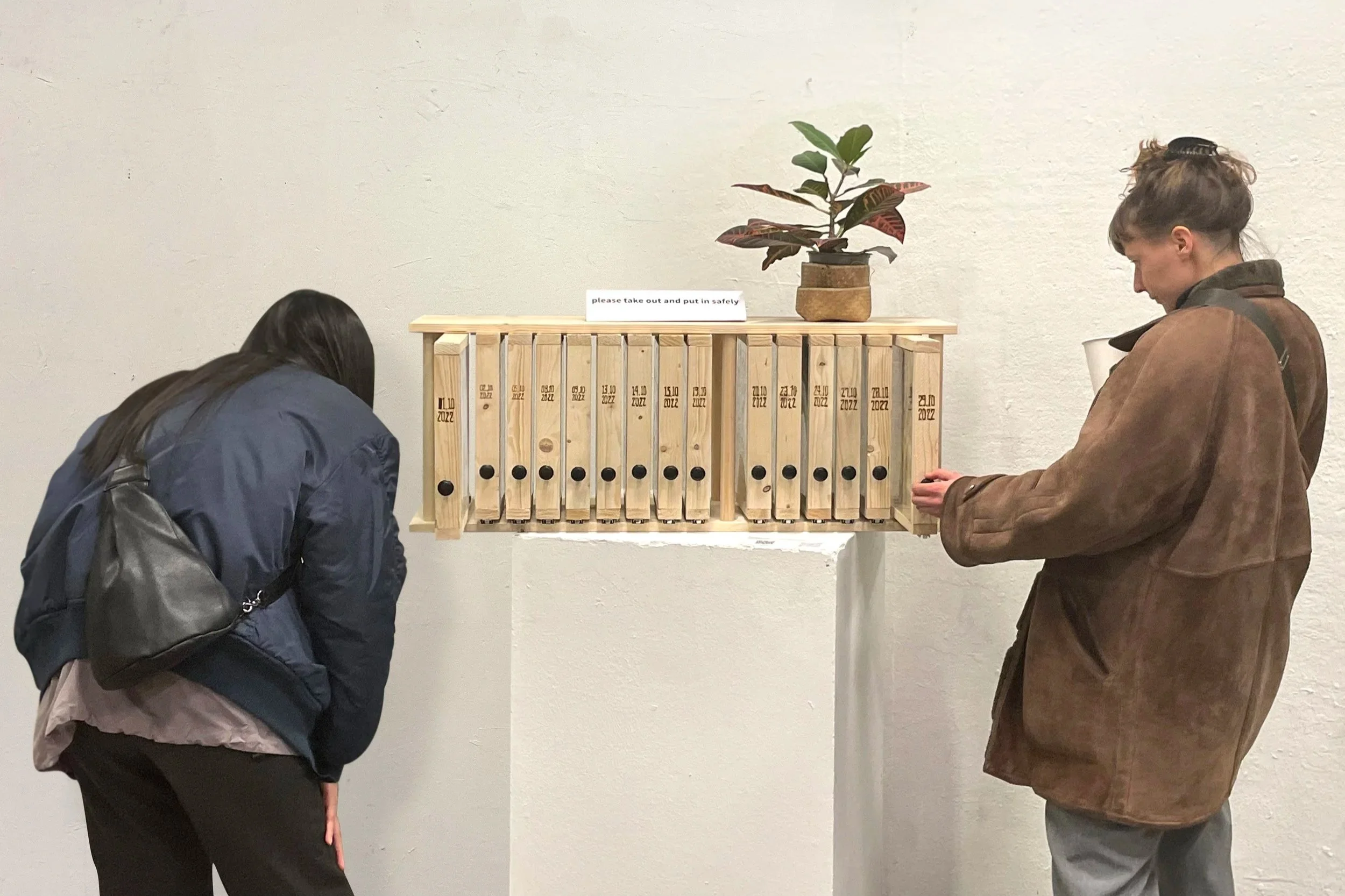 Two women standing near a wooden pickup station with a sign that says 'please take out and put in safely'. A potted plant is placed on top of the wooden pick-up station. One woman is leaning forward and the other is holding a white cup.