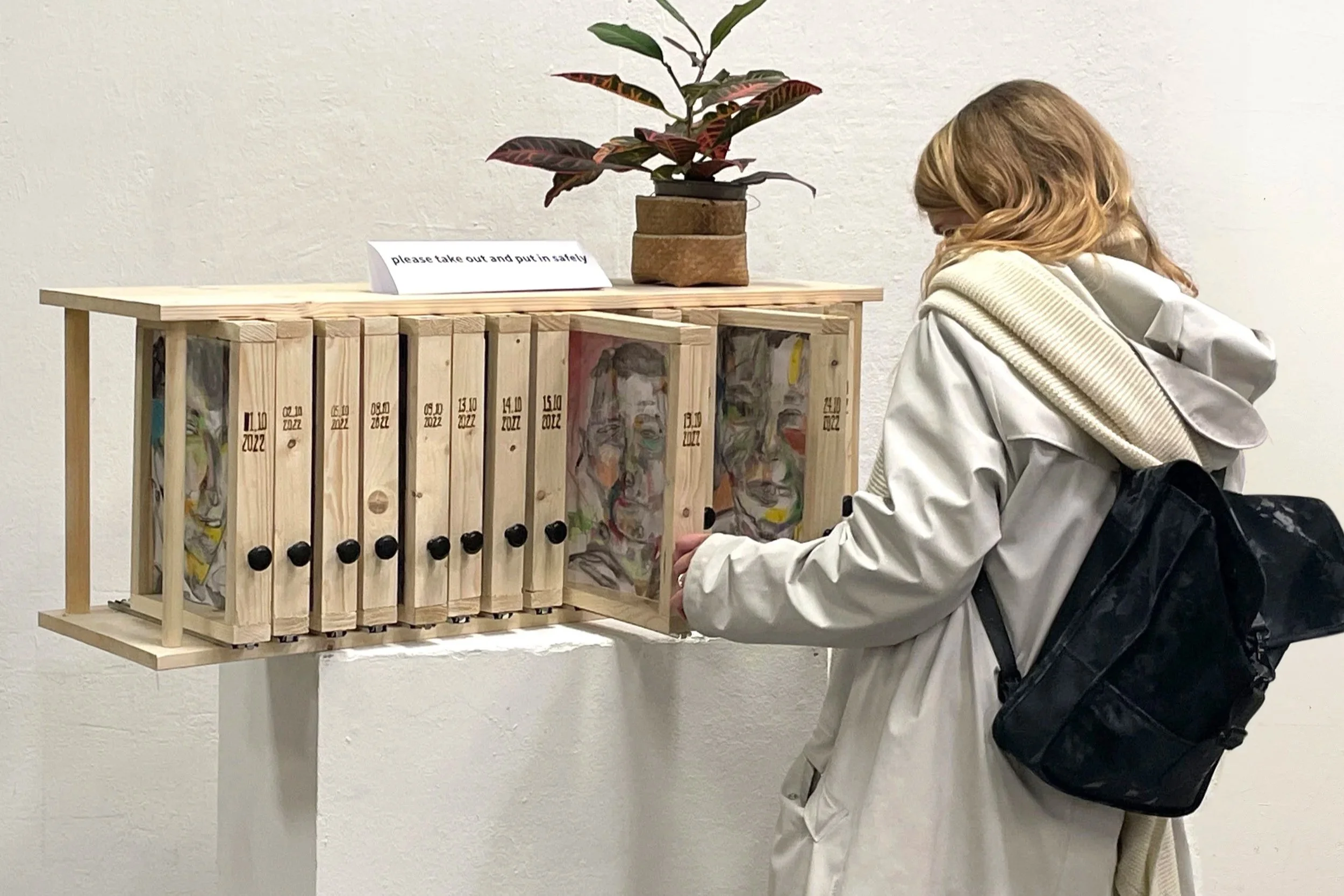 A person with light-colored hair, wearing a white coat and carrying a black backpack, standing at a display case with multiple wooden folders and a potted plant on top. The folders are labeled with dates and have images of a face, and a sign on the table reads, "please take out and put in safety."
