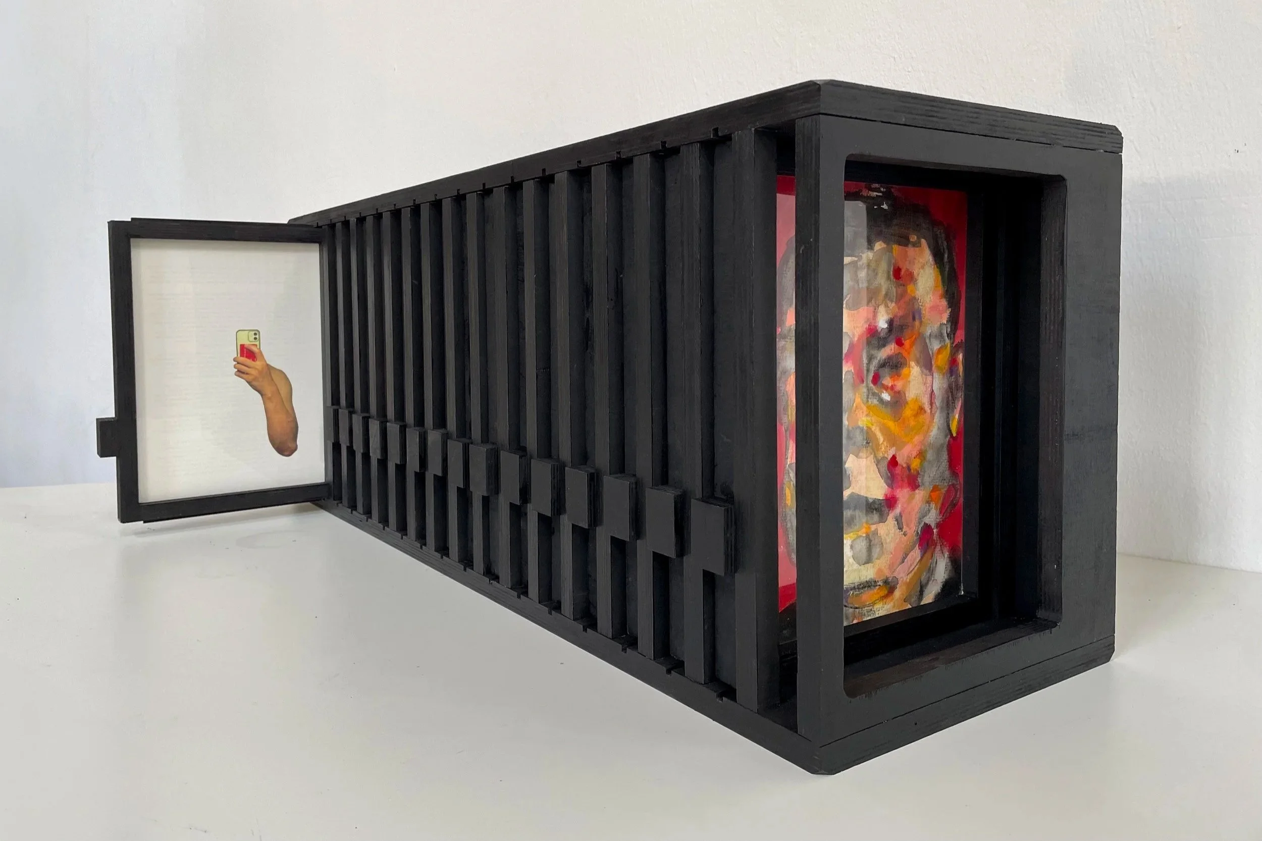 A black wooden box with a glass front displaying an abstract portrait painting inside, and a small mirror on the left side reflecting a hand holding a phone with a yellow case.