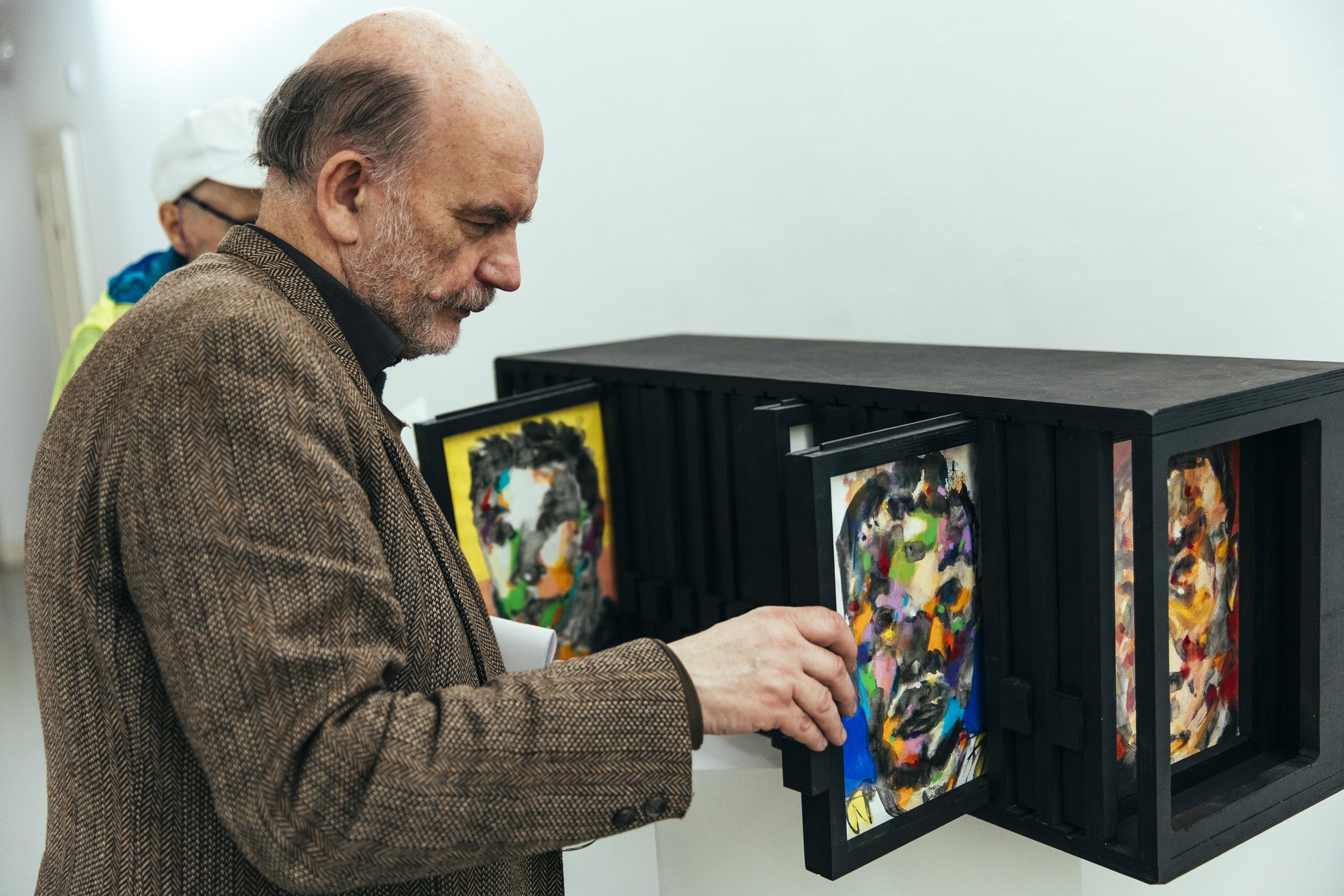 An elderly man with a beard and dressed in a brown jacket looks at colorful abstract portraits of faces displayed in a black frame or box.