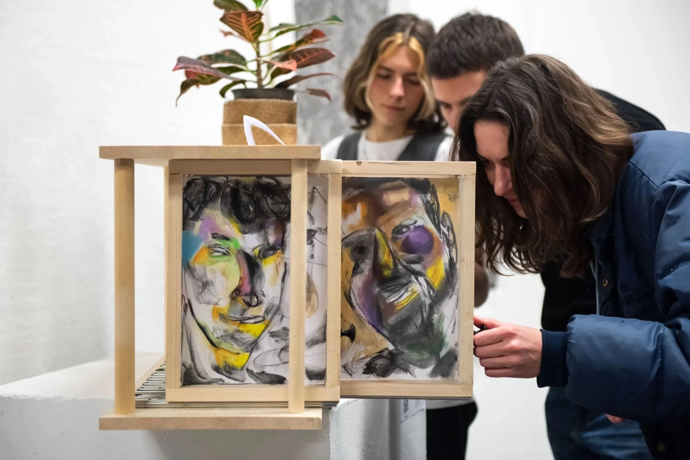 People observing abstract portrait artwork displayed in a wooden frame, with a potted plant in the background.