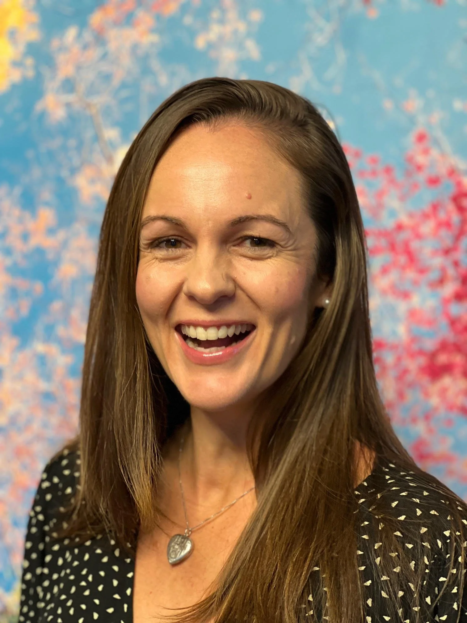 A woman with long brown hair smiling, wearing a black dress with white polka dots and a silver heart-shaped necklace, standing in front of a colorful background with blue and pink hues.