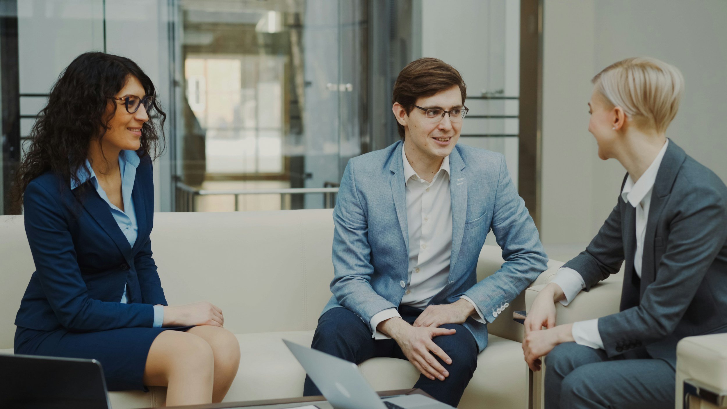 Two people sitting on a couch wearing professional work attire facing another person sitting on a chair. They are in an office setting. They are relaxed and smiling.