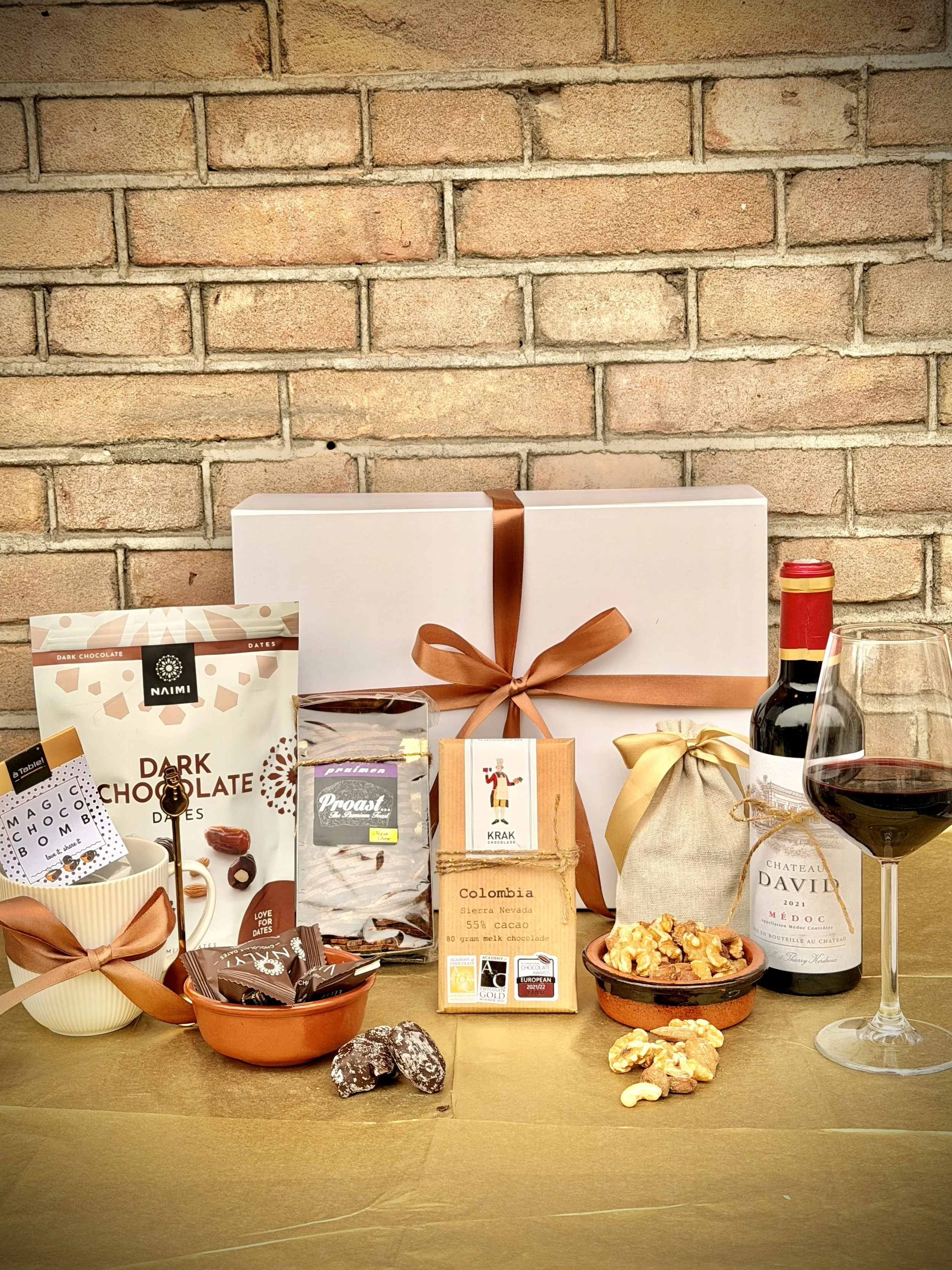 A gift box with a brown ribbon surrounded by chocolates, a bag of dark chocolate dates, a box of coffee, a bottle of wine, a wine glass, and snacks on a wooden table against a brick wall.