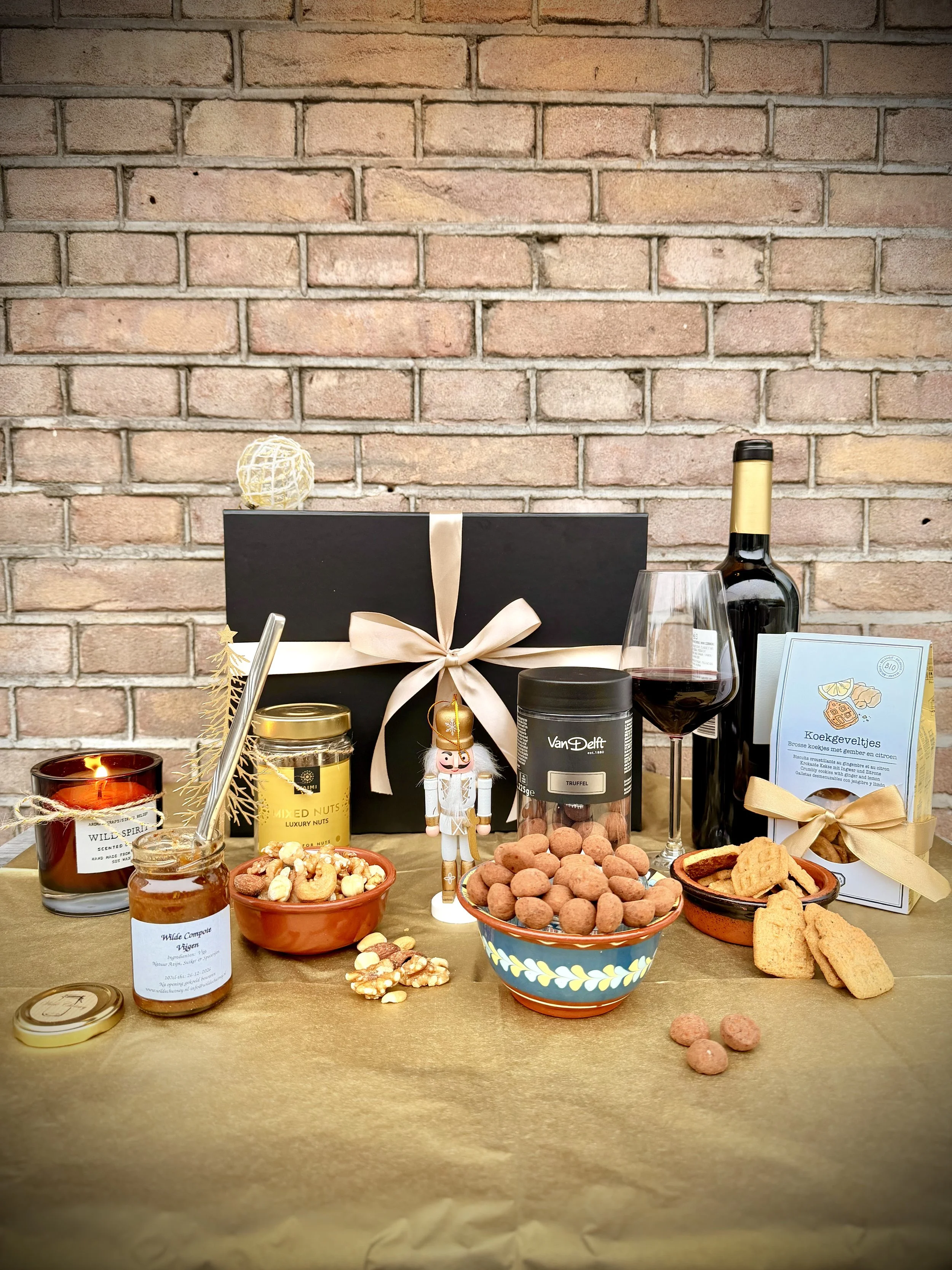 A gift service with snacks and wine, featuring a black box with a pink ribbon, bowls of nuts and cookies, a jar of honey, a nutcracker figure, a bottle of red wine, and a glass of wine, arranged on a gold table against a brick wall background.