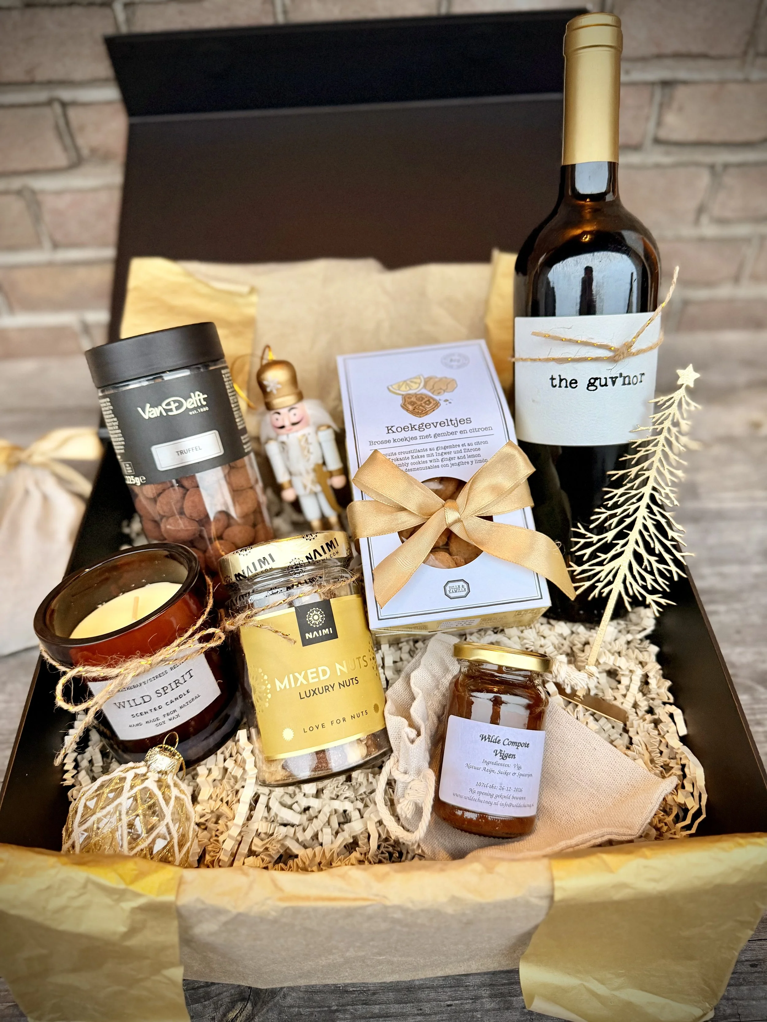 Gift box containing a bottle of red wine with a label 'the guv'nor', a jar of mixed nuts with a gold and black label, a jar of honey, a decorative candle, a small nutcracker, a gold Christmas ornament, and a paper wrapped item, all arranged on crumpl