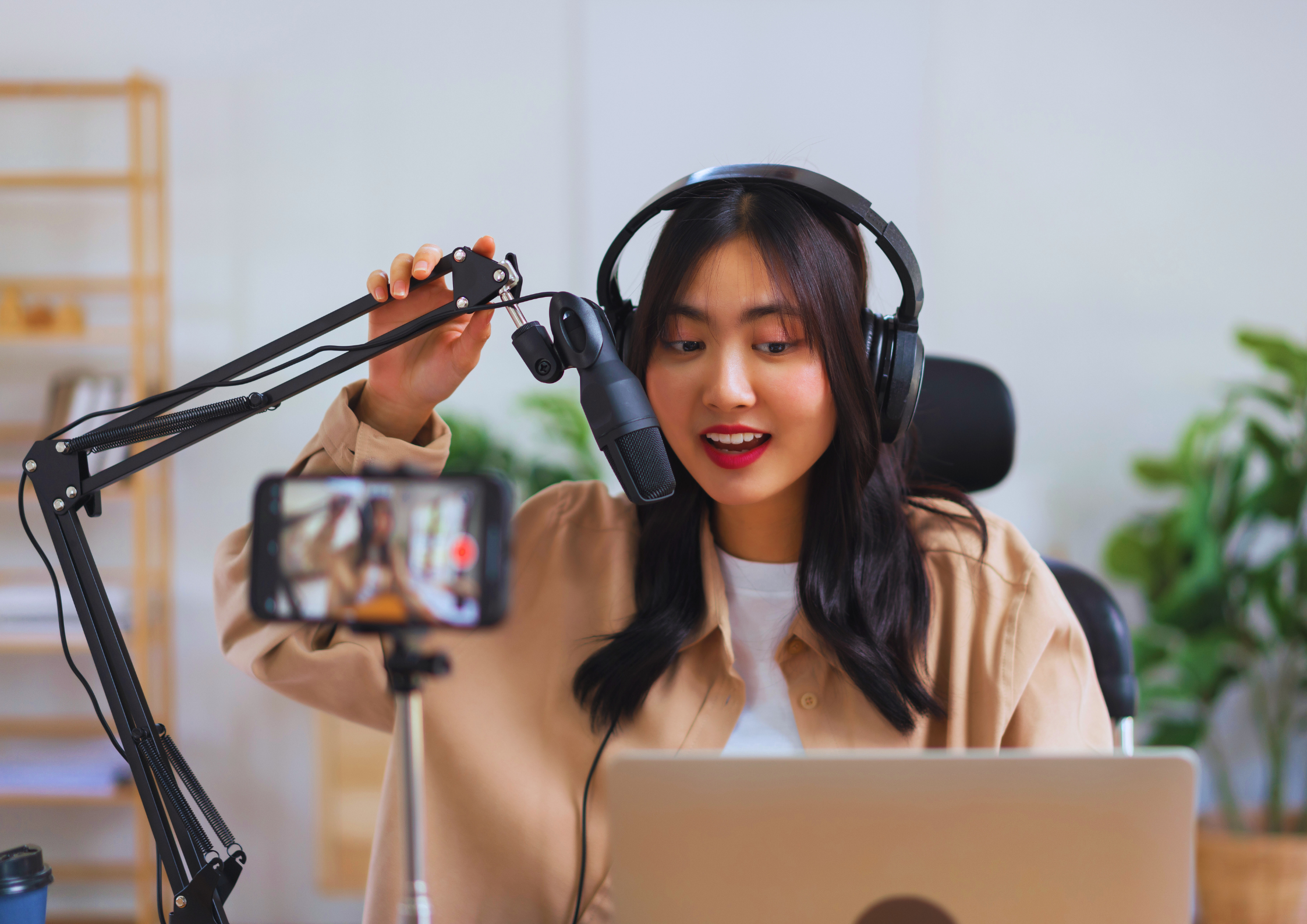 A woman with dark hair wearing headphones and a beige shirt, speaking into a microphone set up on a desk. She is recording a video with a smartphone on a tripod, with a laptop open in front of her. The background has a bookshelf and green plants.