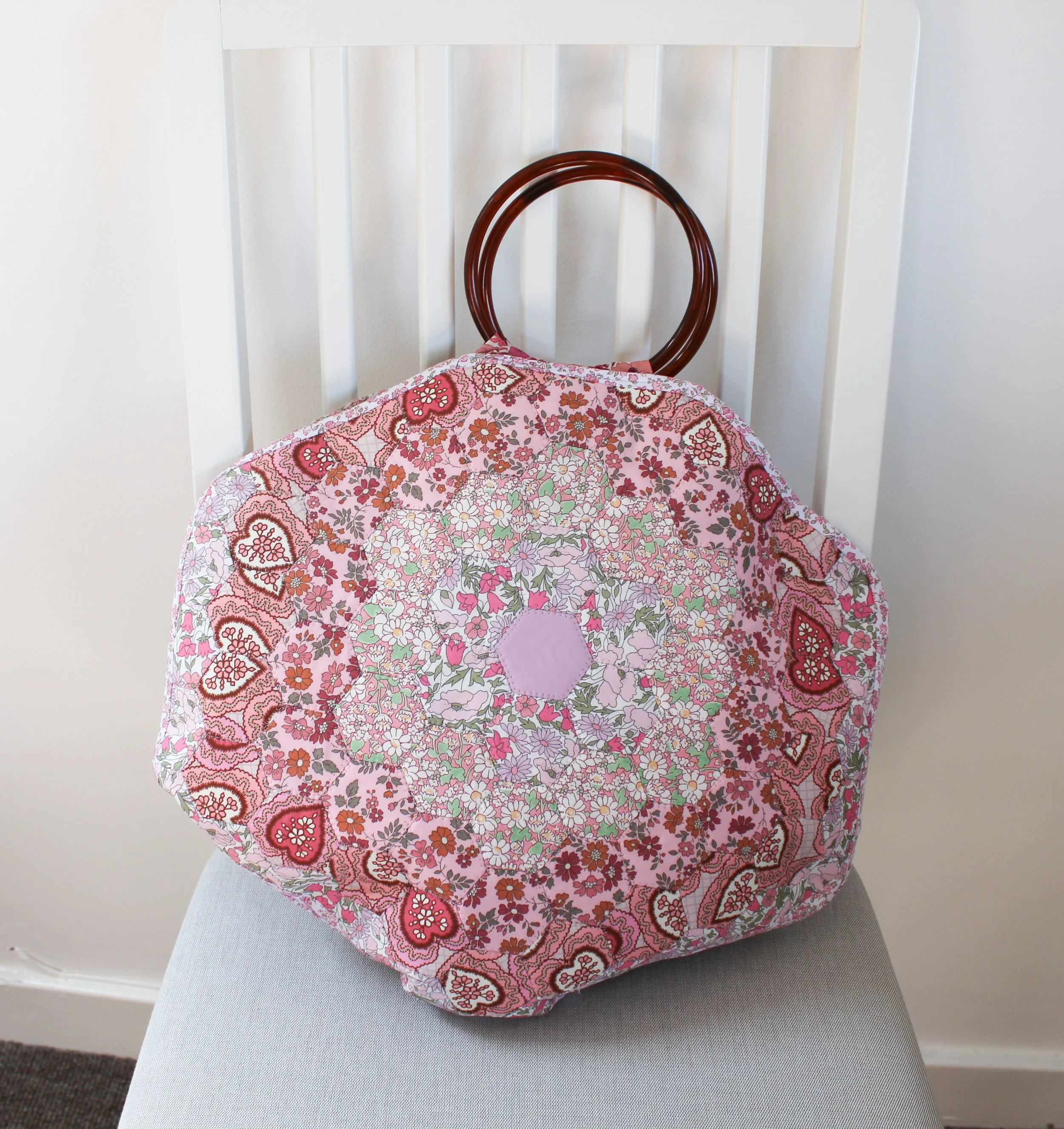 A quilted fabric tote bag with pink, red, and green floral and heart patterns, hanging on a wooden chair with a rounded backrest, placed on a gray upholstered surface in front of a white wall with vertical paneling.