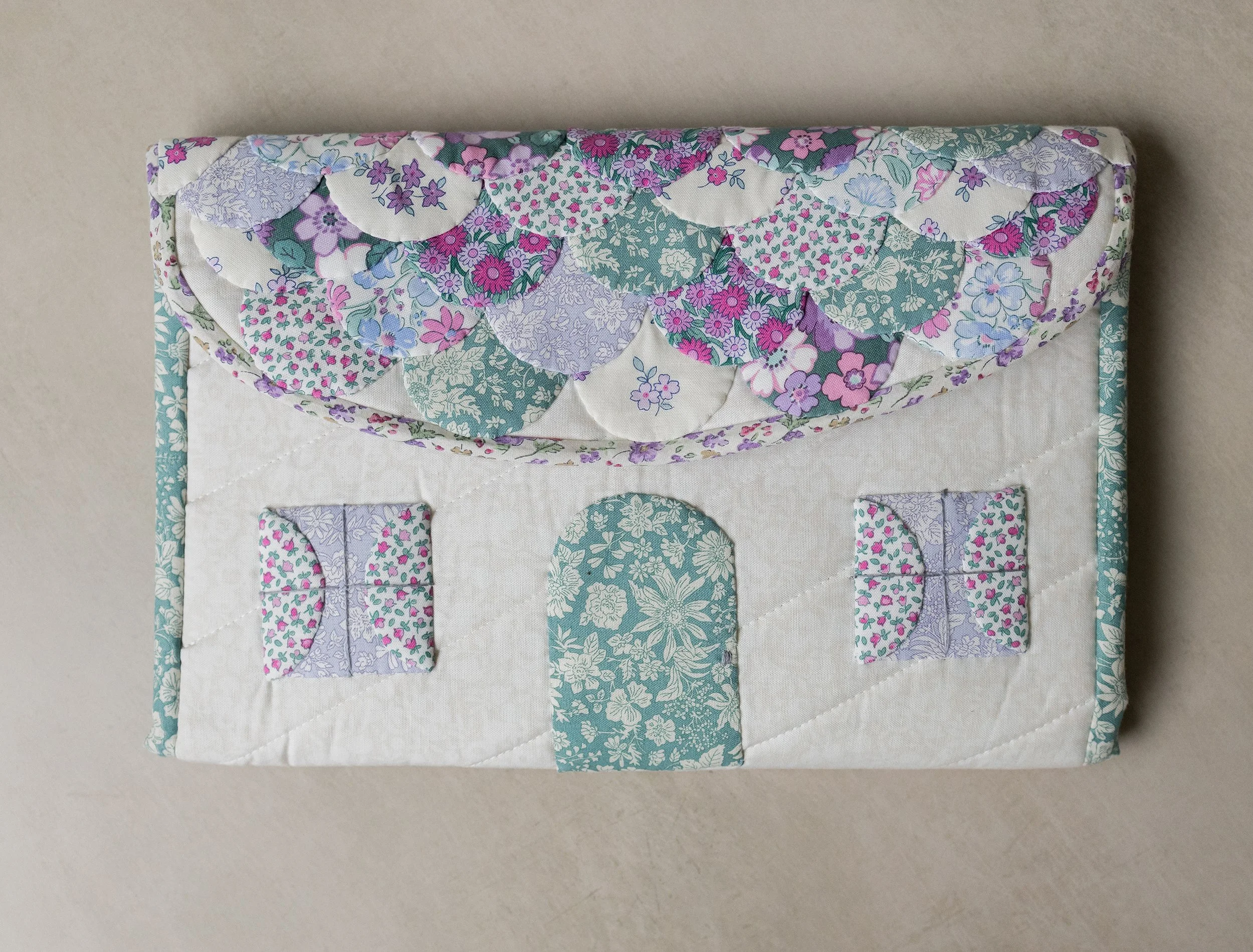 A quilted fabric wall hanging depicting a house with a floral patterned roof, door, and windows, all stitched onto a white background.