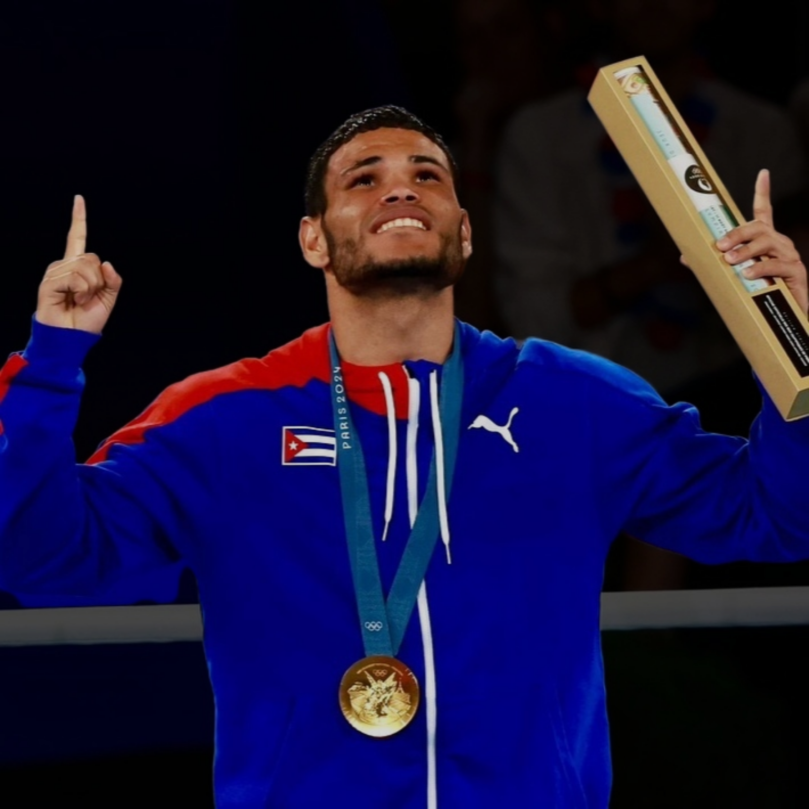 ERISLANDY ALVAREZ boxeur cubain portant une médaille d'or aux Jeux Olympiques, tenant un trophée, regarde vers le haut, vêtu d'un sweat bleu avec le drapeau cubain et un logo Puma.