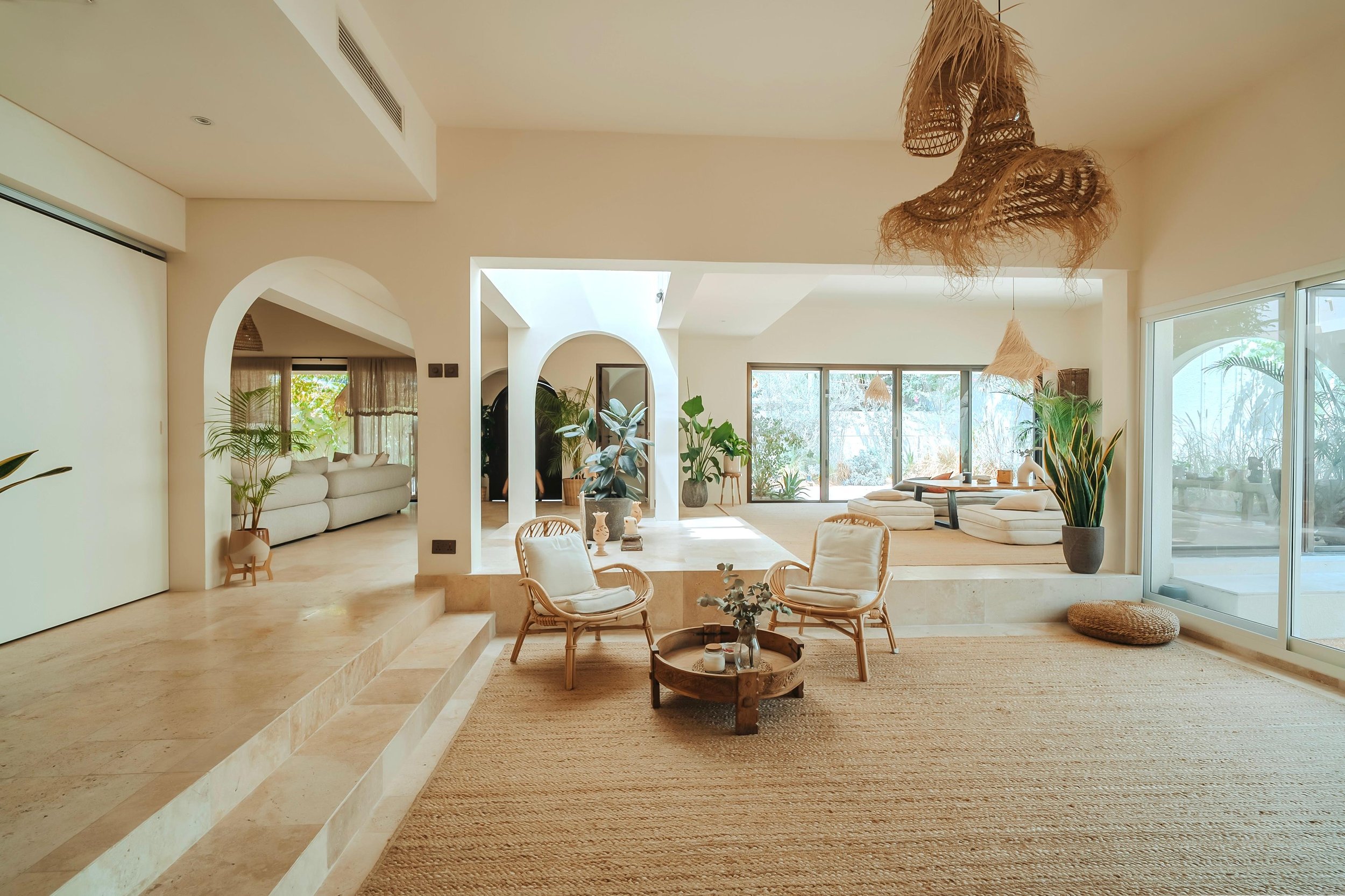 Bright living room with natural light, beige and white decor, rattan chairs, large plants, and sliding glass doors with outdoor view.