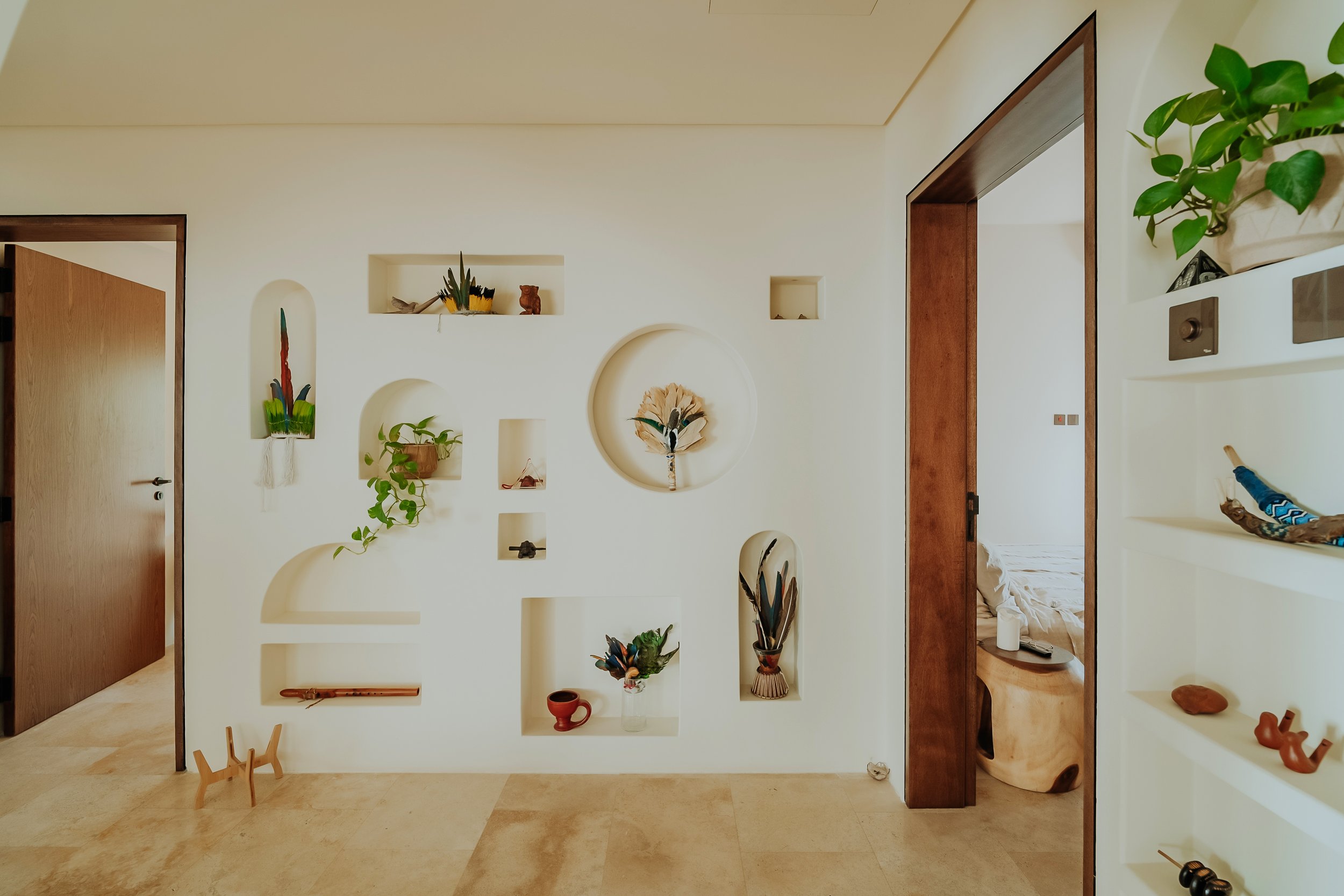 A minimalist interior living space with a white decorative wall featuring various built-in niches displaying plants, ceramics, and art pieces. There's an open doorway revealing a bedroom with a bed and side table, and shelves on the right with small decorative objects. The floor is tiled with beige stone, and there's a small wooden stool in the corner.