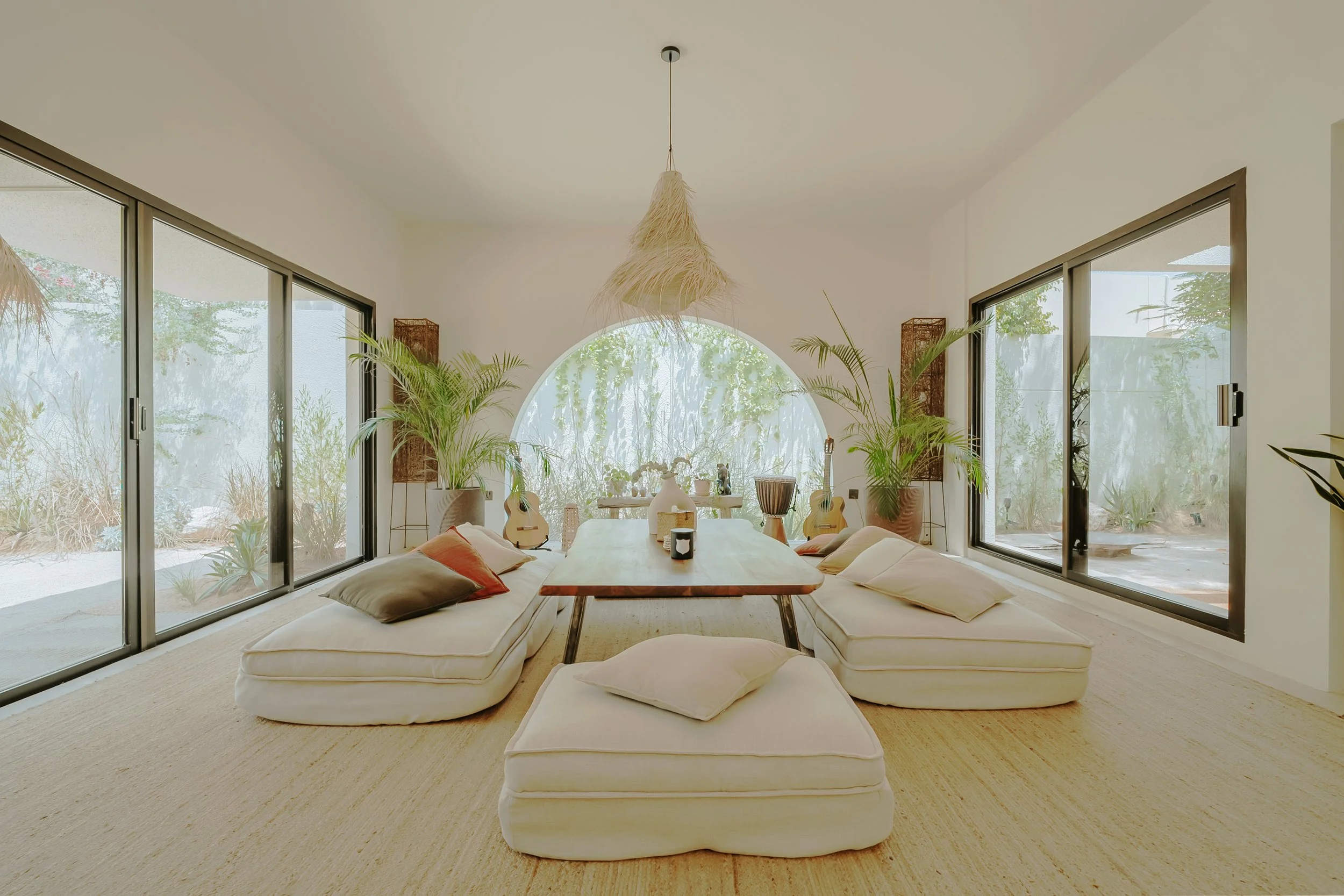 Bright living room with large floor-to-ceiling windows, a central wooden table, bean bag chairs with pillows, indoor plants, guitars, and decorative items.