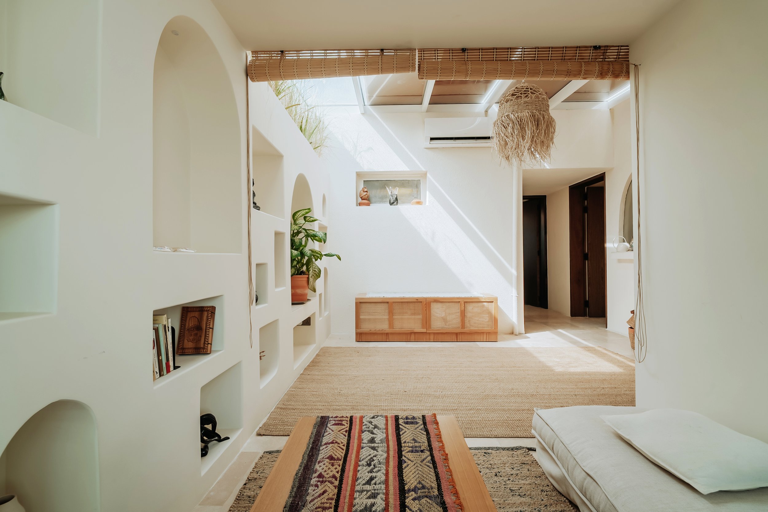 Bright living room with white walls, built-in shelves, a potted plant, a woven rug, a low wooden bench, and a ceiling light. Sunlight streams in from a skylight, illuminating the space.