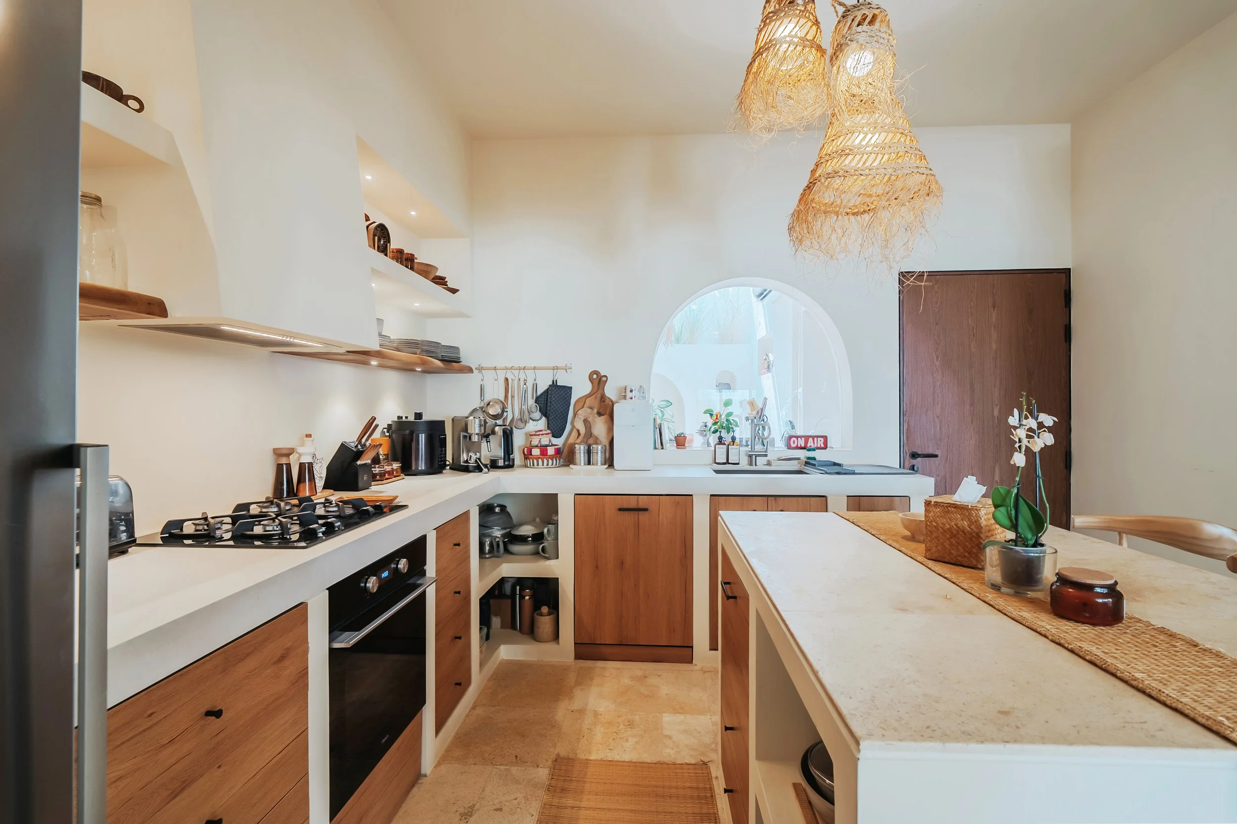 Bright kitchen with white walls, wooden cabinets, open shelving with dishes, a gas stove, and a sink under a round window; decor includes a orchid plant, woven light fixtures, and various kitchen appliances.
