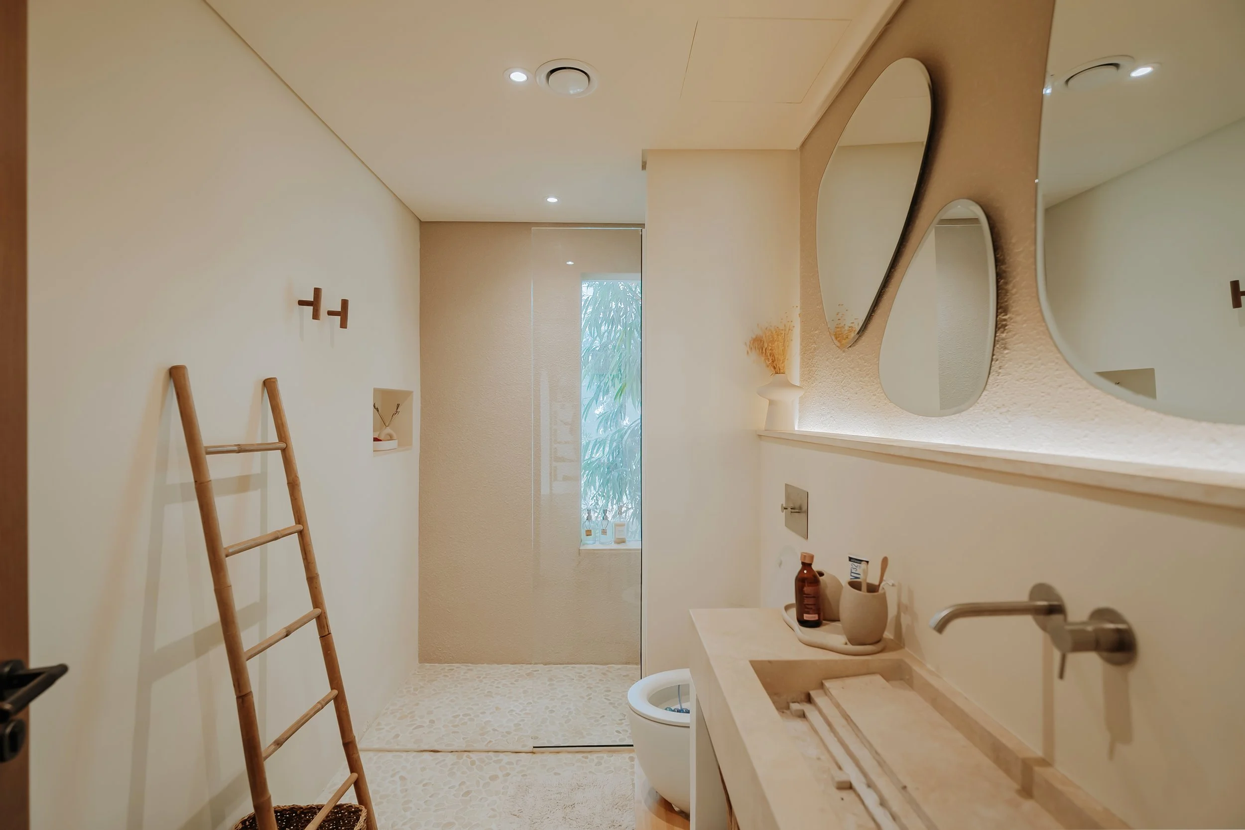 Minimalist bathroom with beige walls, wooden ladder, bathroom sink with toiletries, wall-mounted mirror, and a walk-in shower at the back with a textured beige floor.