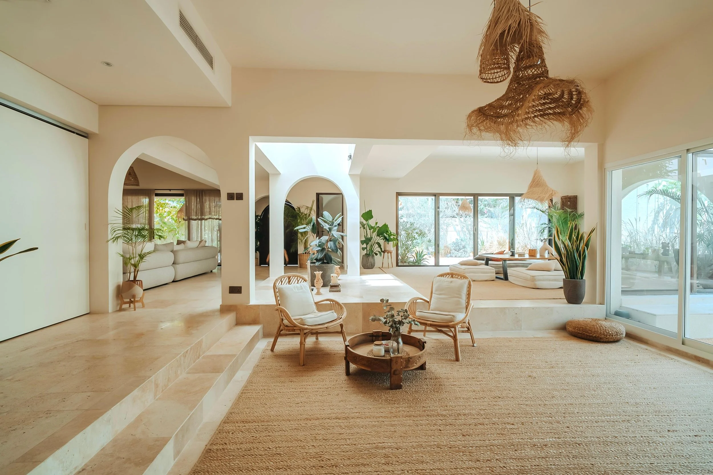 Bright, modern living room with large windows, neutral tones, indoor plants, rattan furniture, and woven light fixtures.