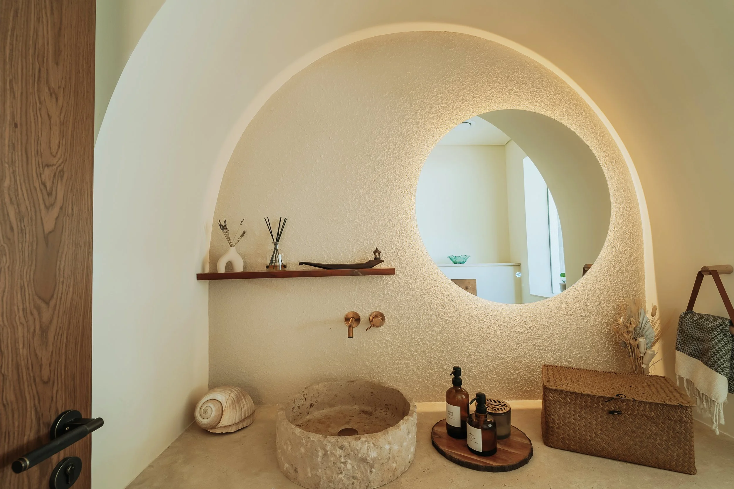Interior of a minimalistic bathroom featuring a stone sink, a round mirror, a wooden shelf with decorative items, and a woven basket on the floor.