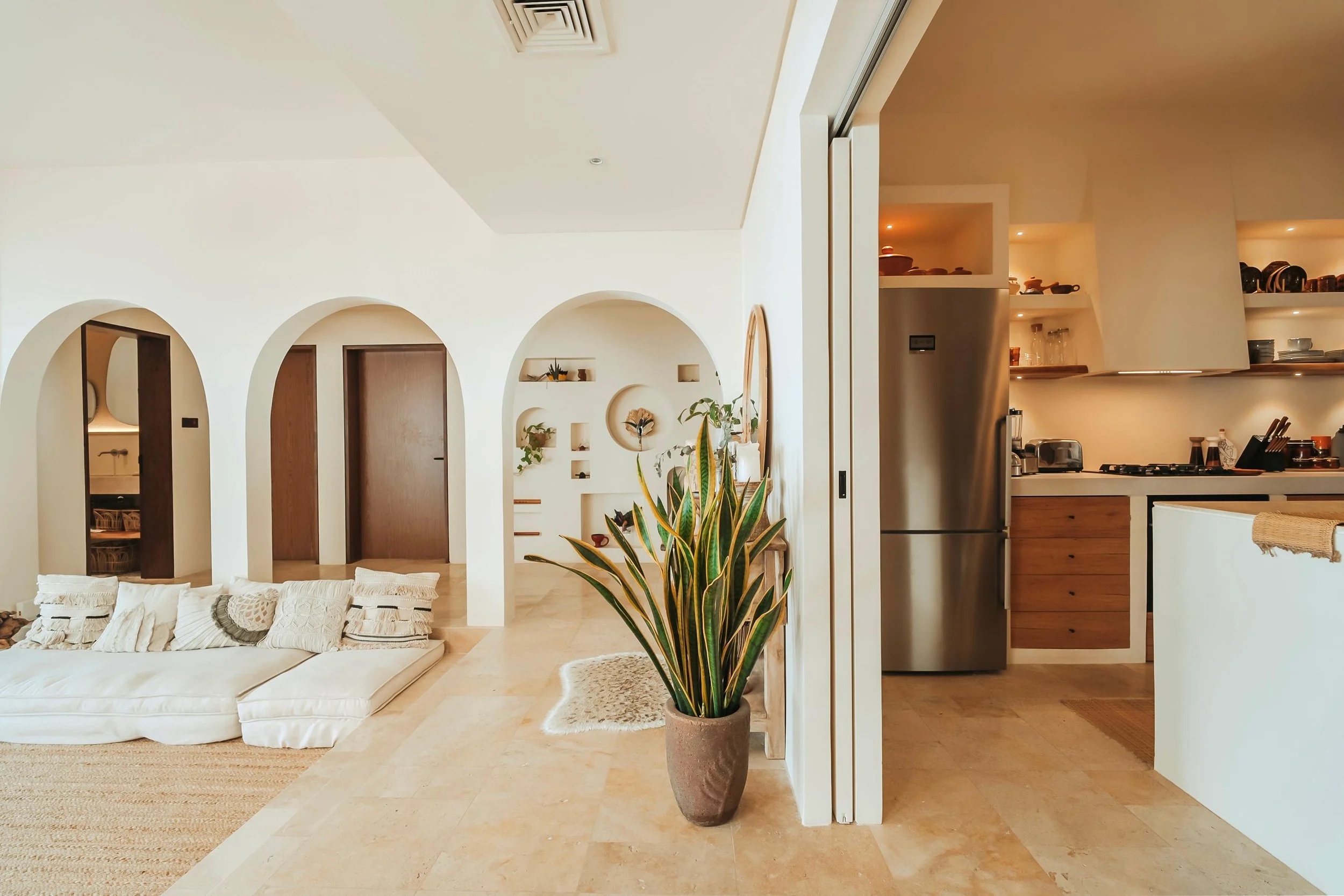 Interior view of a modern, minimalistic home with white walls, arched doorways, a beige tiled floor, a large potted plant, a white sofa with decorative pillows, and an open kitchen with wooden cabinets and stainless steel appliances.