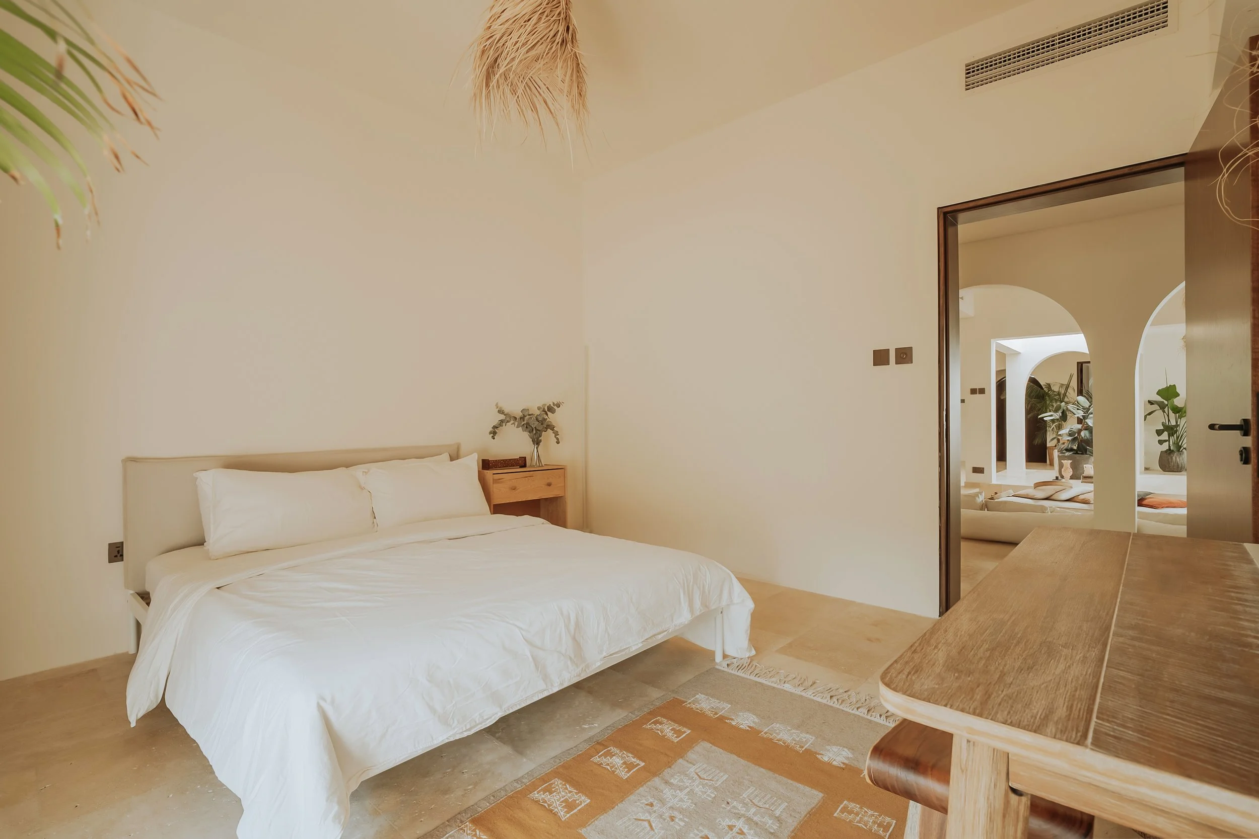 A bedroom with a white bed and pillows, wooden side table with a vase of flowers, beige walls, and a view into another room with plants and seating areas.