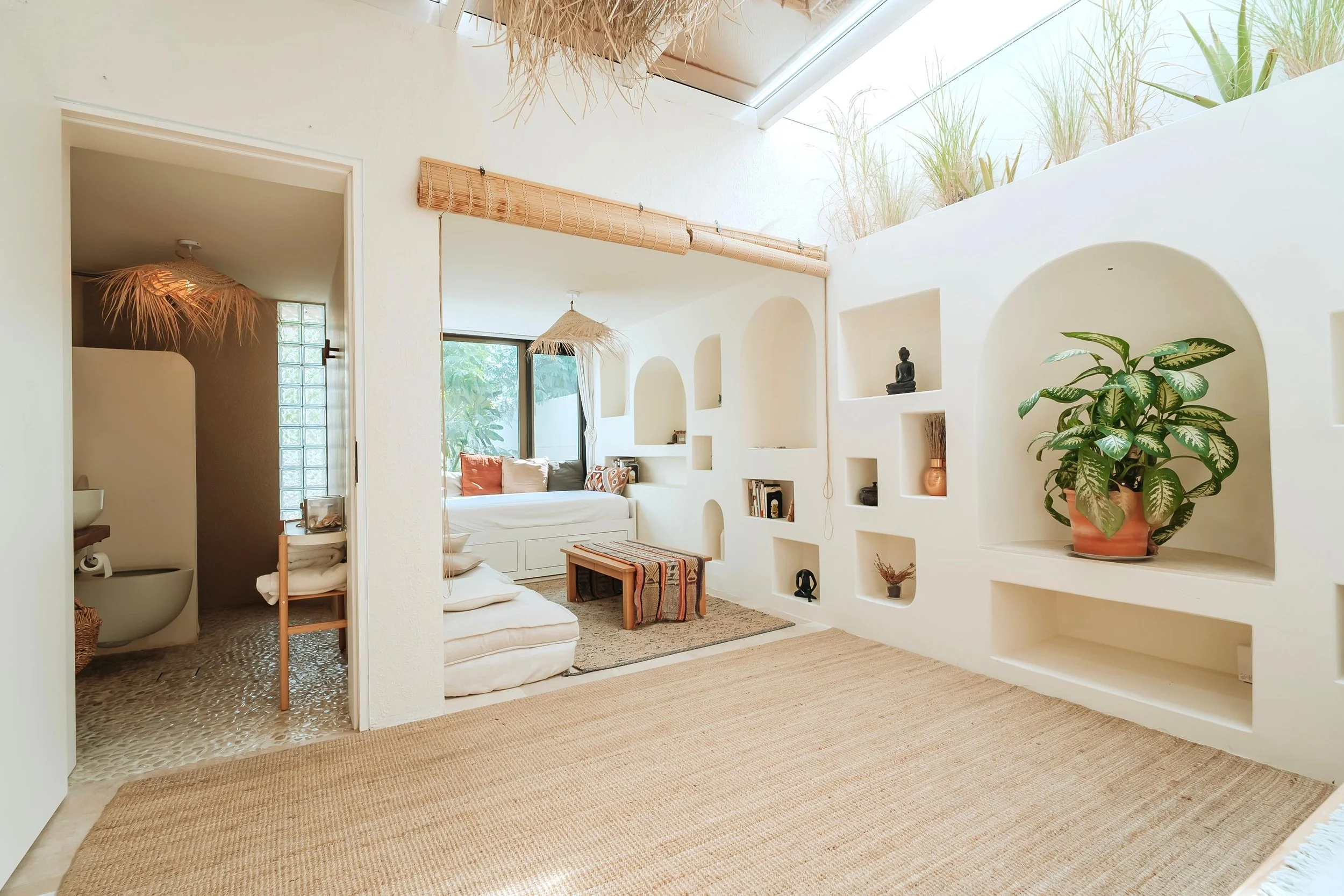 Bright, airy living space with white walls, built-in shelves, and a large potted plant. There are two sitting areas with cushions, a window seat with pillows, and natural decor including rugs, baskets, and bamboo accents. Skylights and a glass door provide natural light.