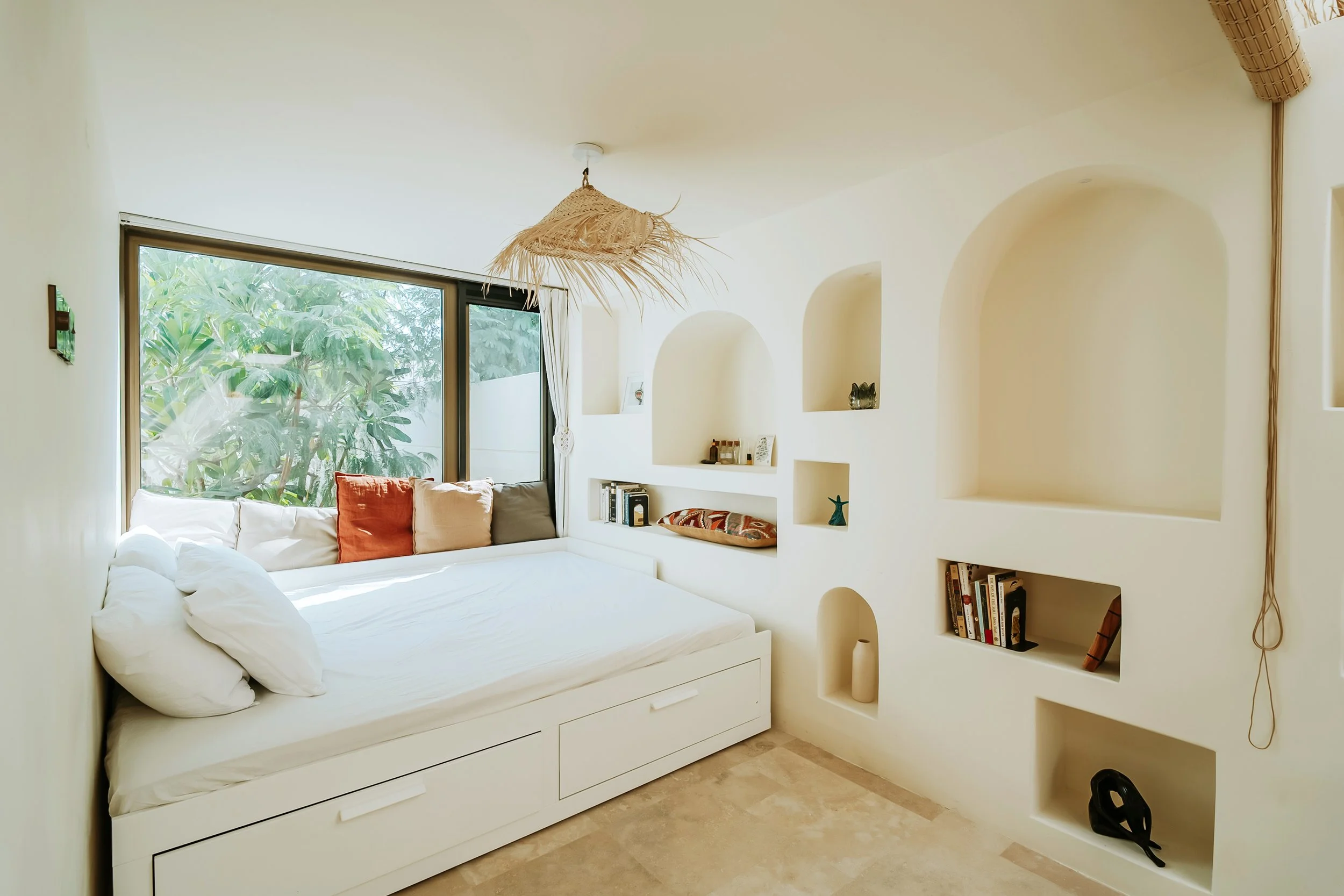 A cozy bedroom with a built-in bed next to a large window with lush greenery outside. The wall features multiple recessed wall niches with decorative items and books, and a woven pendant light hangs from the ceiling.