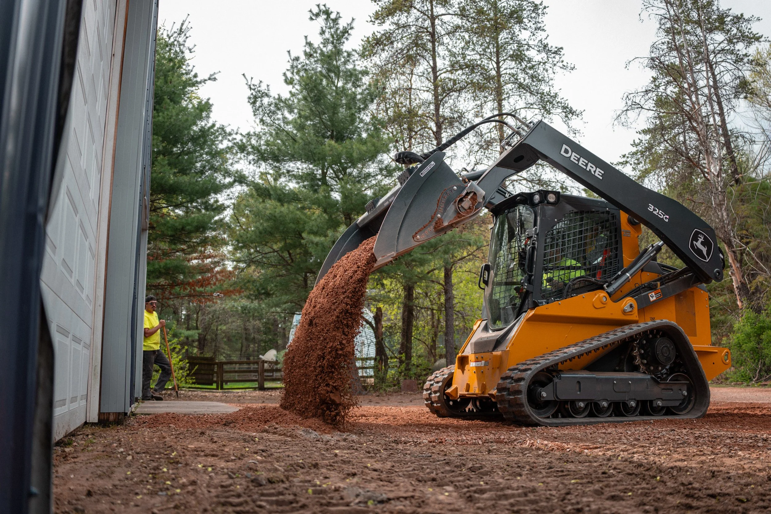 A John Deere compact excavator is used to move dirt or mulch onto a pile in a backyard with tall trees. A man wearing a bright yellow shirt is standing nearby, observing the work.