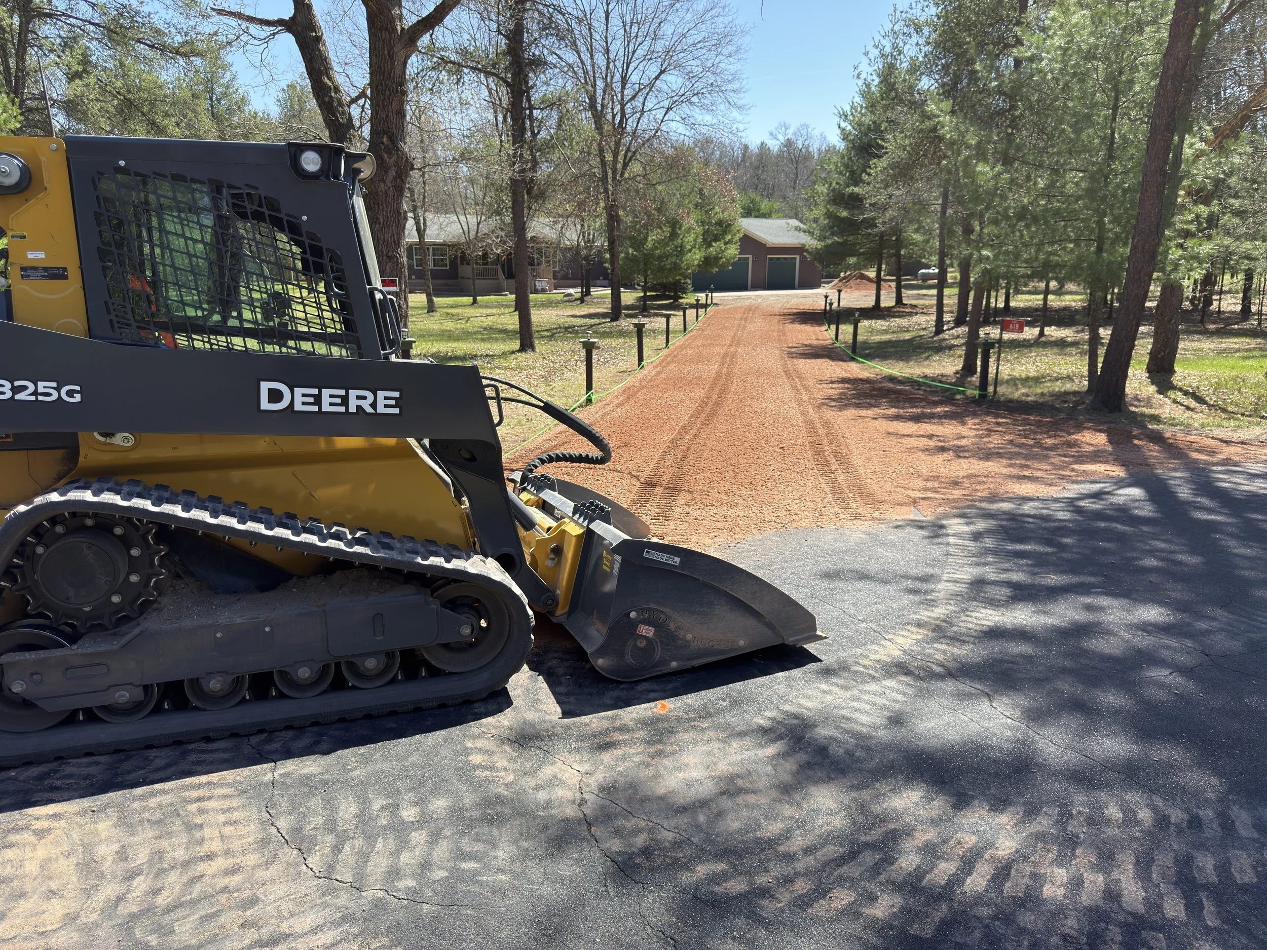A yellow John Deere compact construction vehicle on a paved surface with a background of trees and a house. The vehicle appears to be for landscaping or construction work.