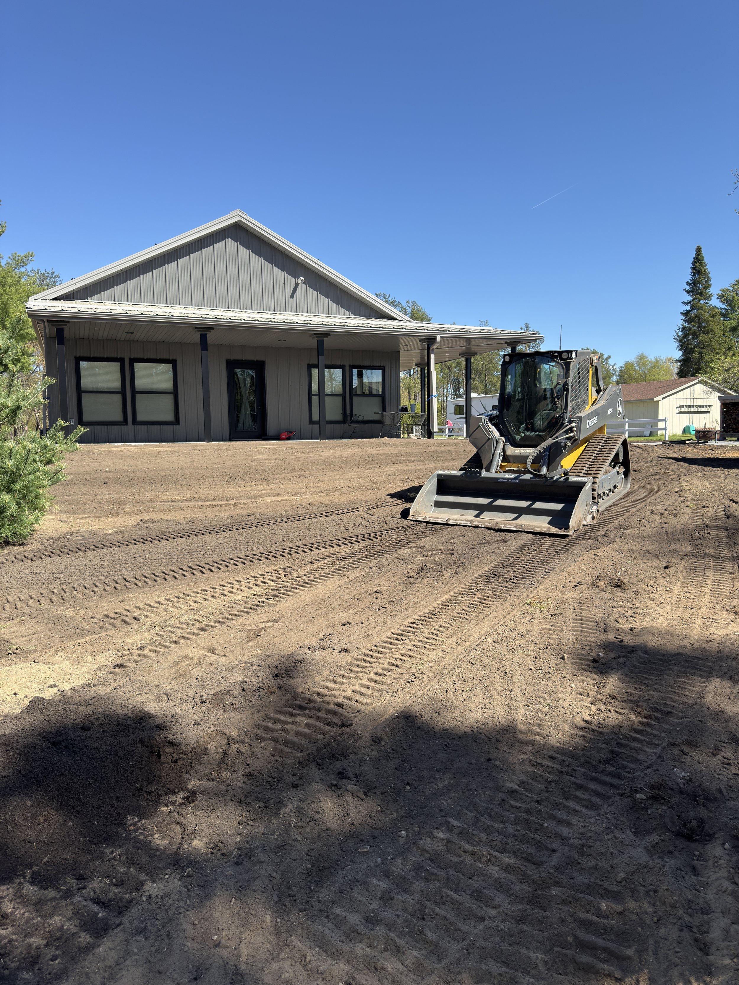 A construction site with a New Holland skid steer loader on freshly tilled dirt in front of a gray house with a wide porch and black-framed windows, and a clear blue sky overhead.