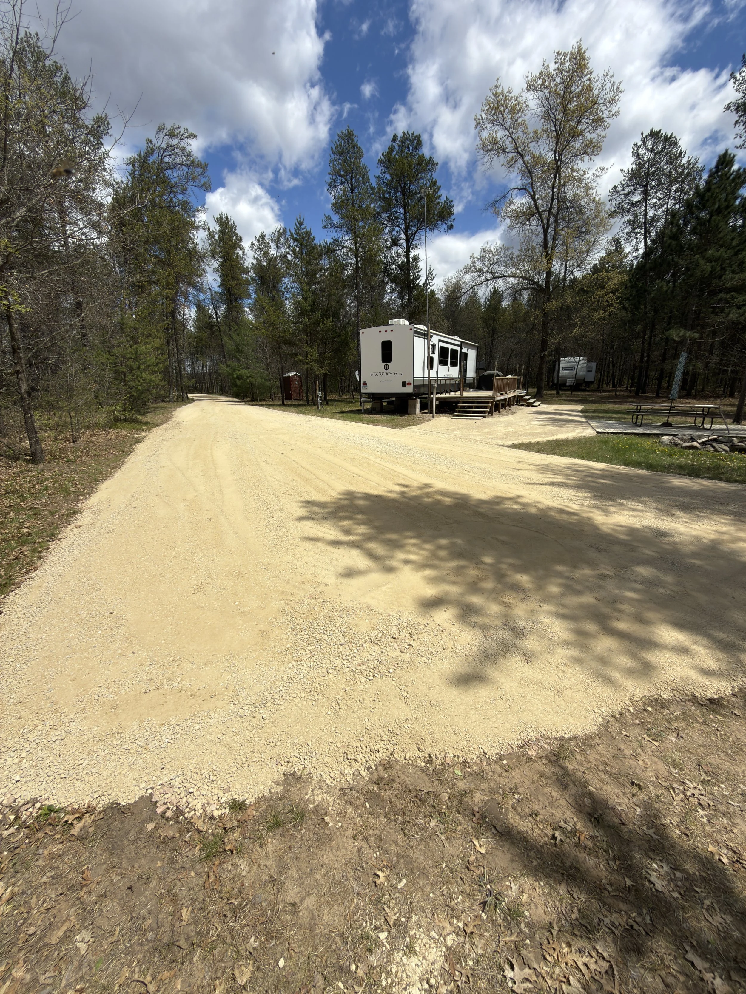 A dirt road curves through a wooded campground with trees, picnic tables, and RVs under a partly cloudy sky.