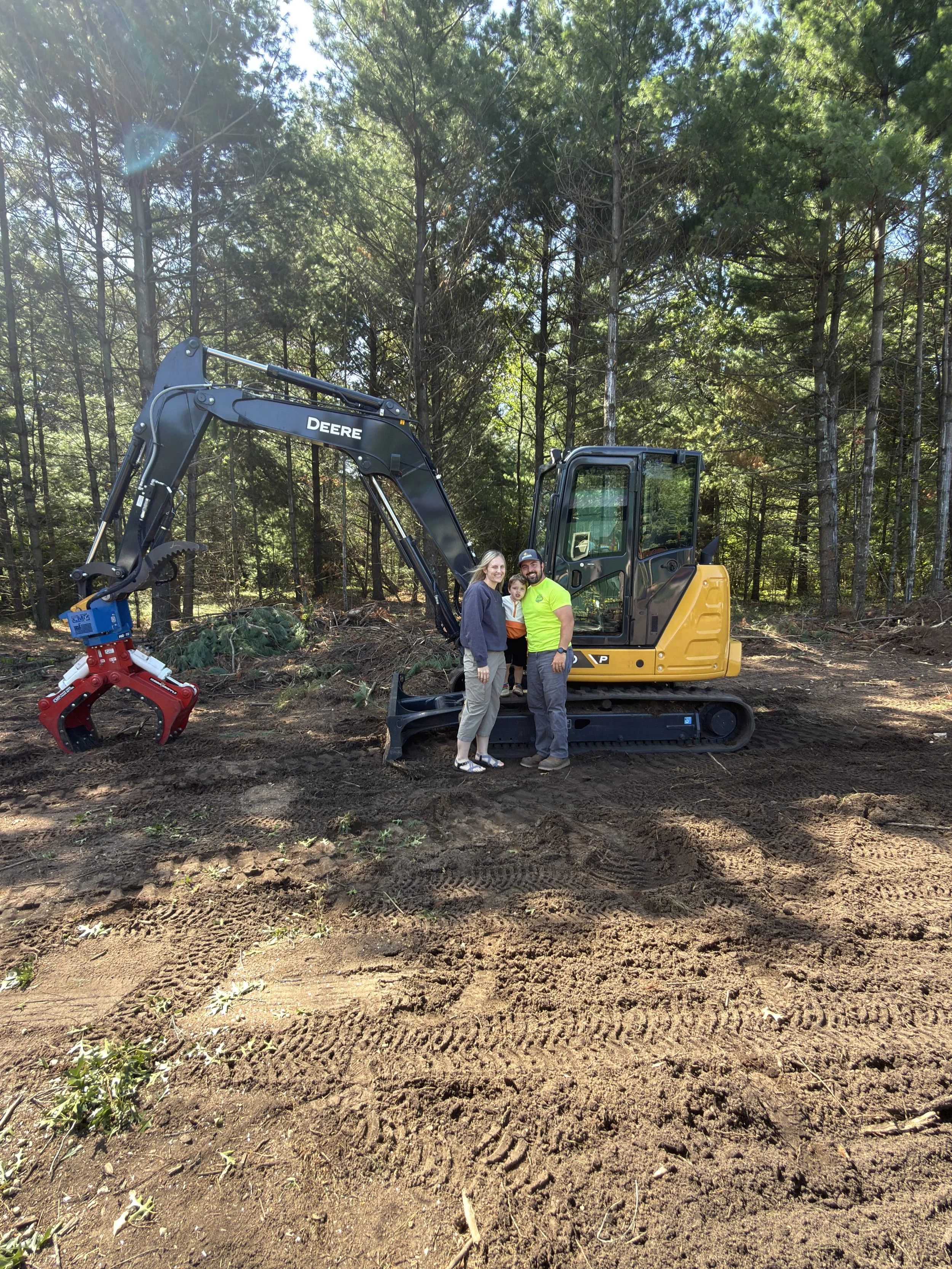 Two children in a wooded outdoor area, one boy in a colorful shirt and shorts on a platform with electronic equipment, and a girl partially visible, observing a small construction vehicle lifting a tree.