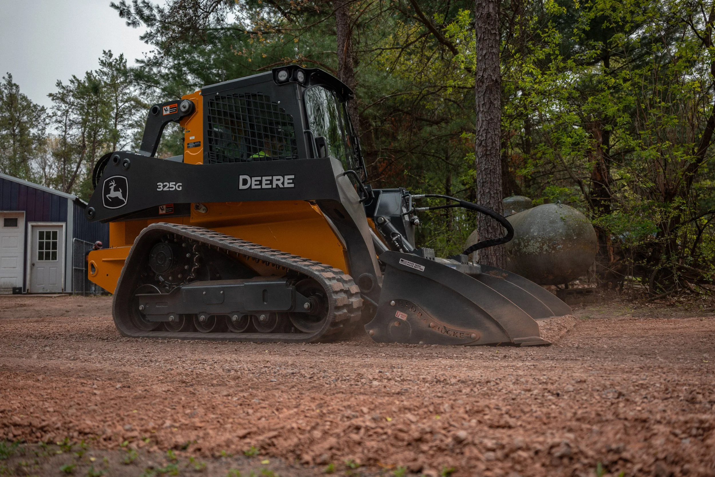 A John Deere compact track loader with a front bucket attachment working on a dirt ground in a wooded area with trees in the background.