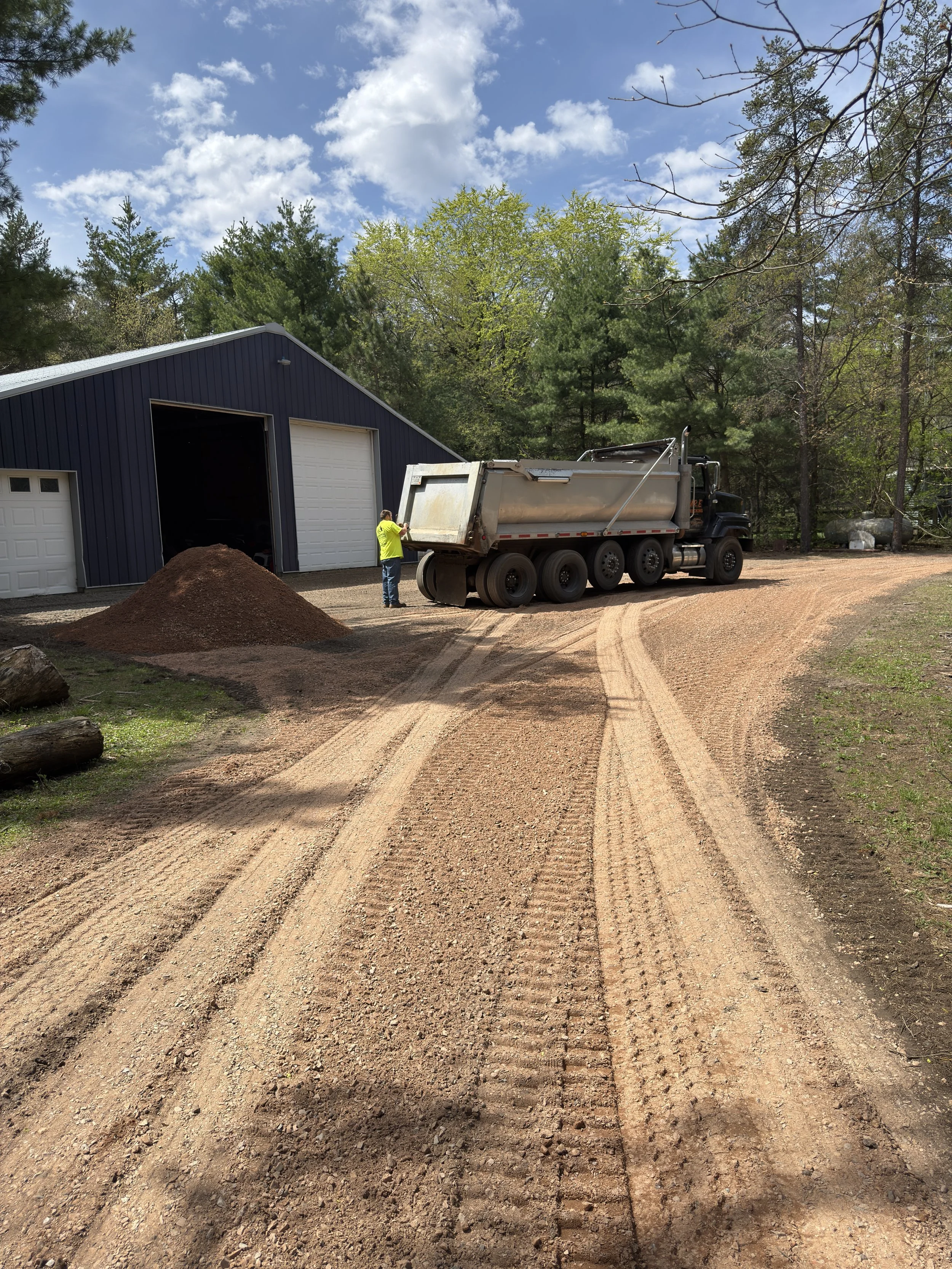 A person in a yellow shirt is standing next to a large construction dump truck on a dirt road, near a blue metal building with three garage doors. There is a pile of dirt in the foreground and trees and a partly cloudy sky in the background.