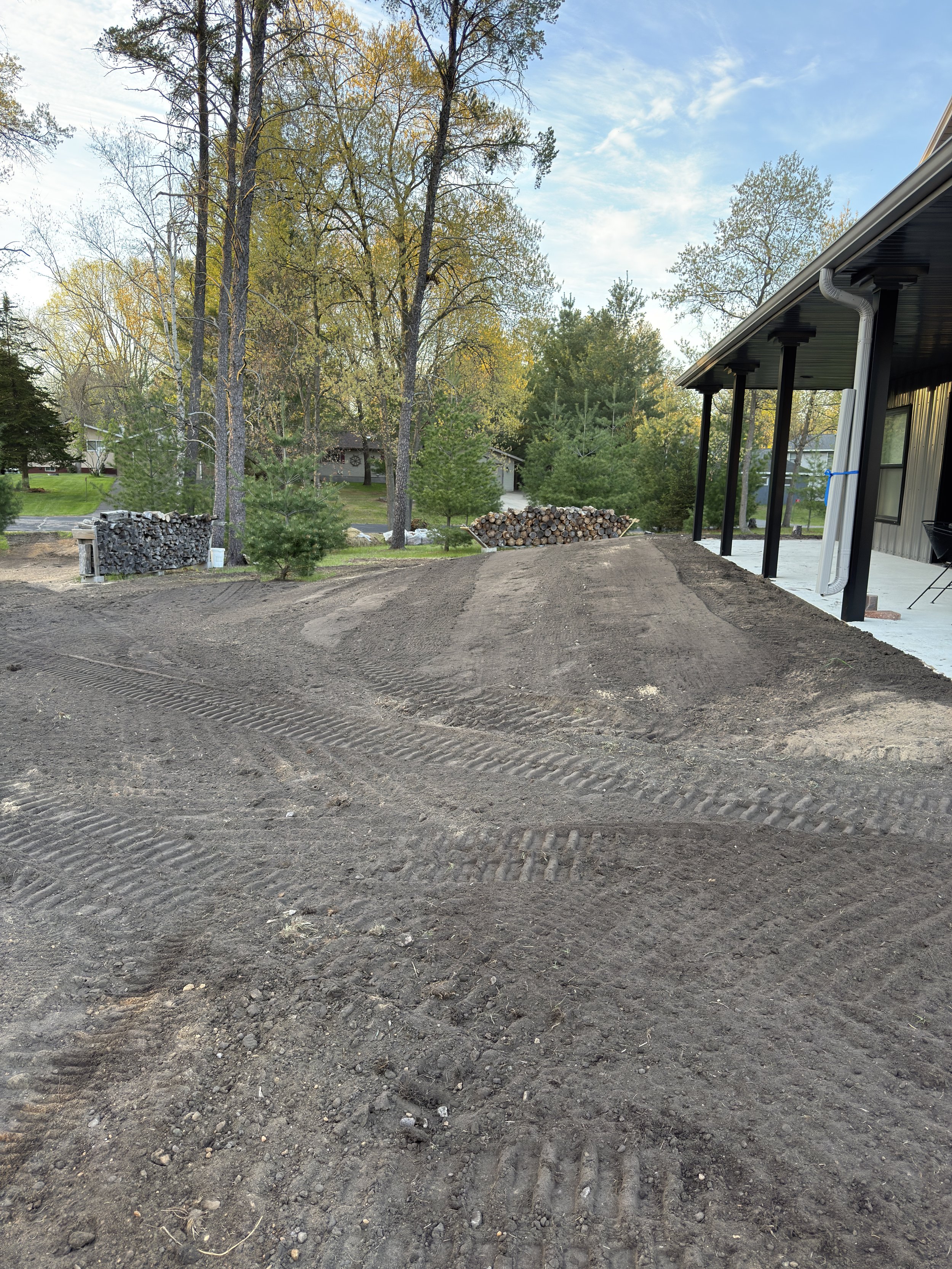 View of a construction site showing fresh dirt with tire tracks, a stack of firewood, and a house with a covered porch, trees, and a partly cloudy sky in the background.