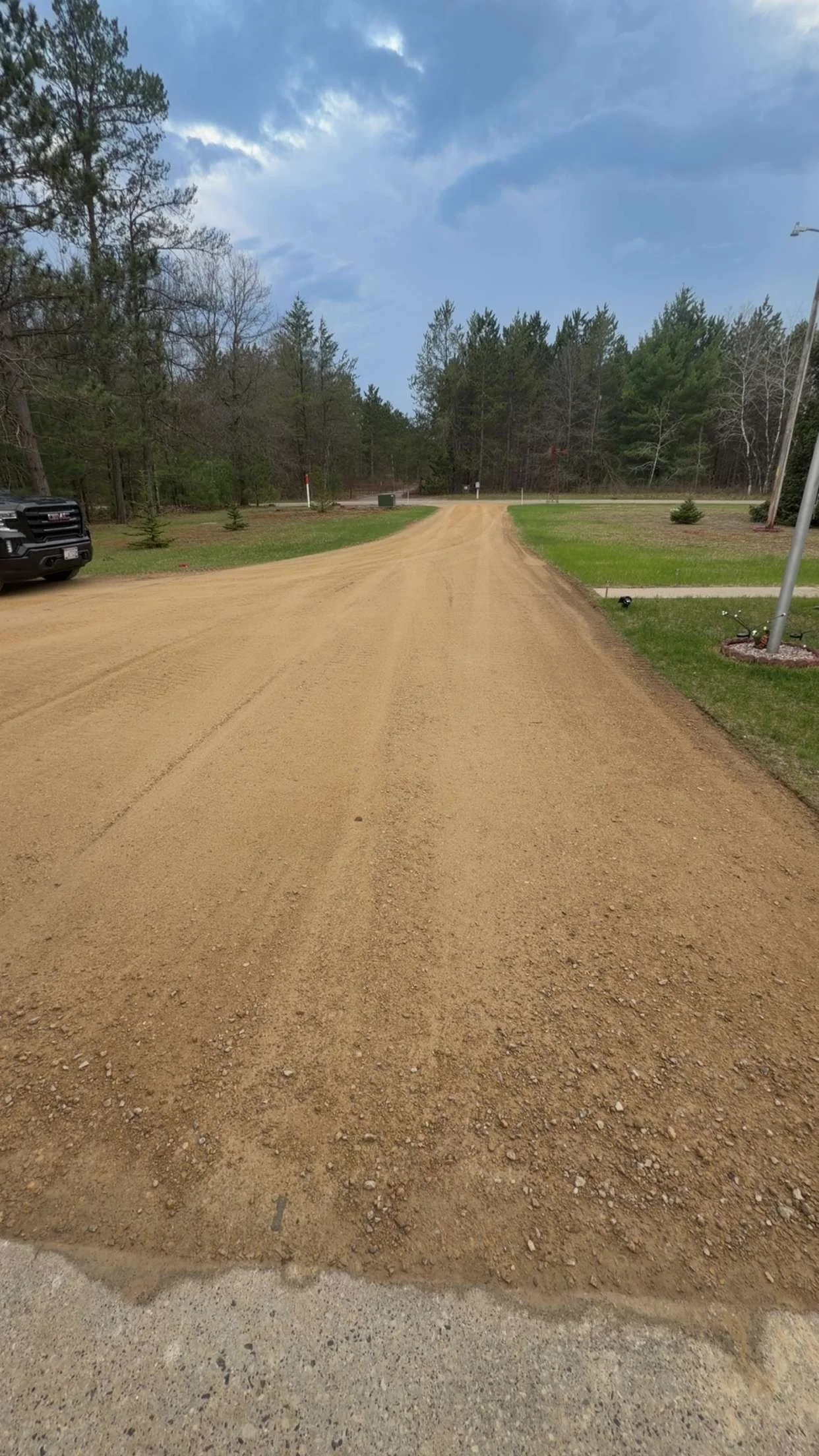 A dirt road with a black vehicle parked on the left side, trees in the background, a cloudy sky, lamp posts, and small plants near the sidewalk.