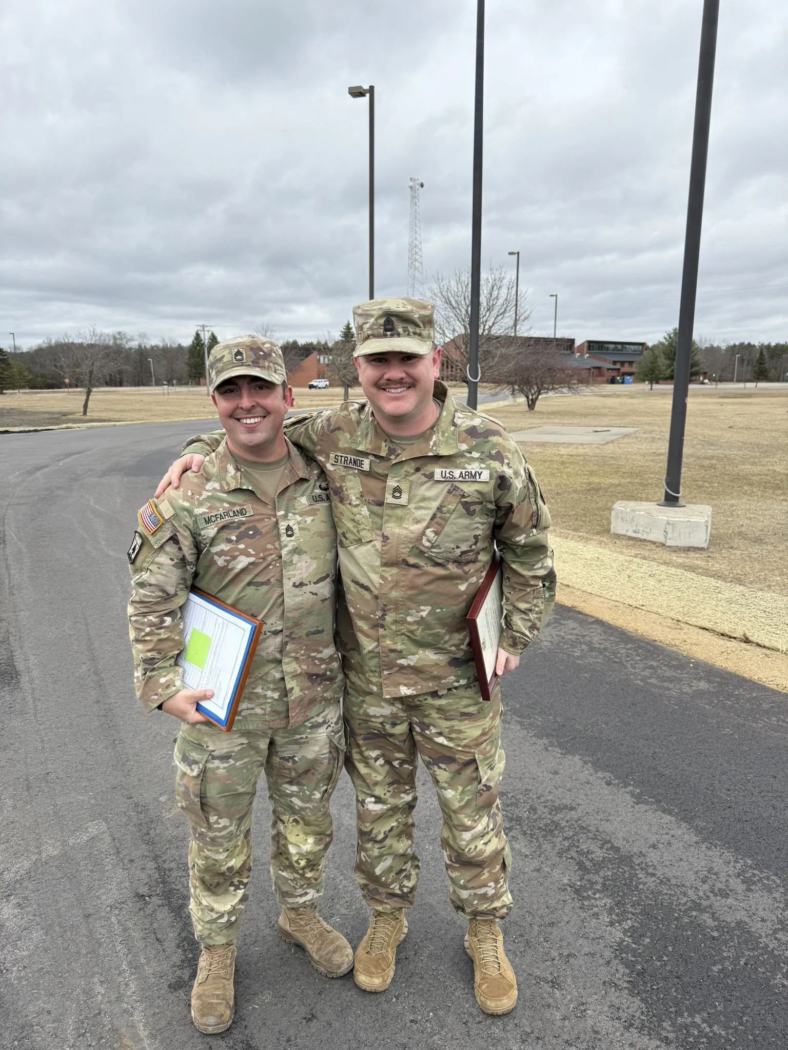 Two soldiers in camouflage military uniforms standing outdoors on a paved area, smiling and holding documents, with a grassy field, trees, and a building in the background, under a cloudy sky.