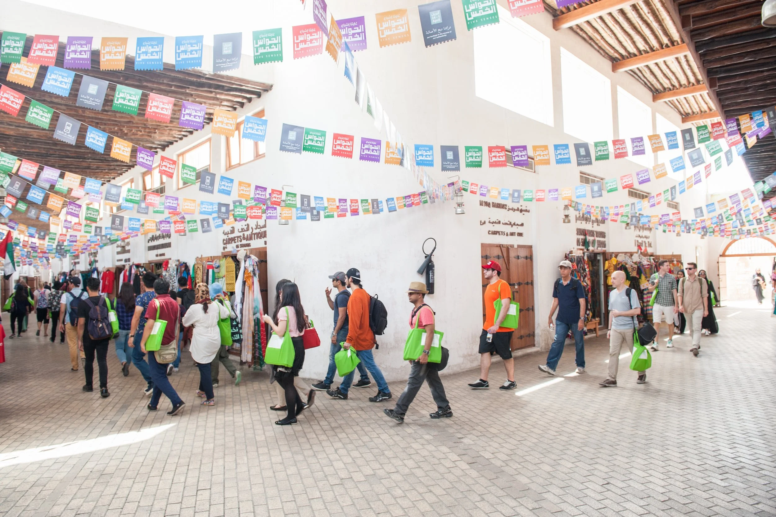 People shopping and walking in a busy indoor marketplace with colorful banners hanging from the ceiling.