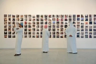 Three people in thobes and head coverings viewing a large wall with numerous small photographs in an art gallery.