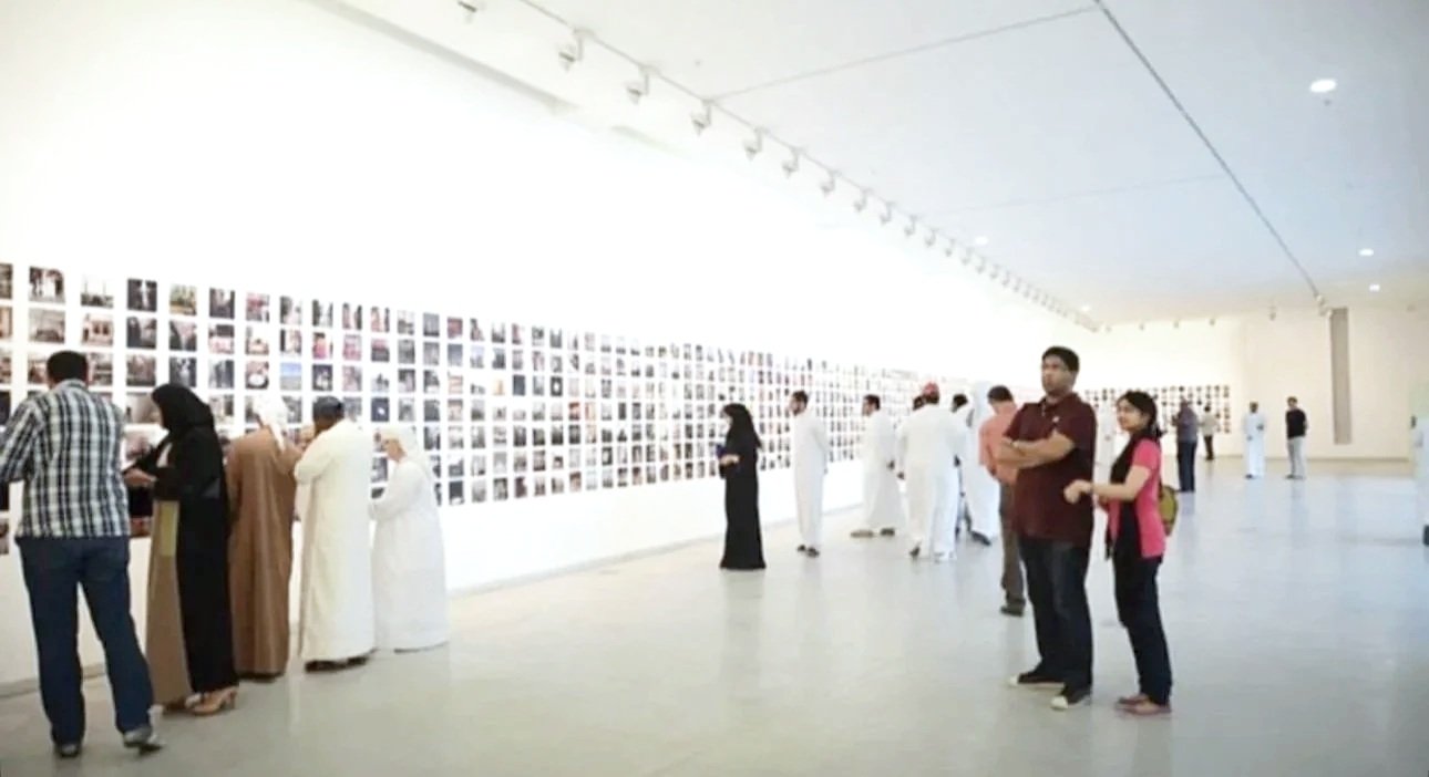 People viewing an art exhibition with a wall of photographs in a spacious gallery.