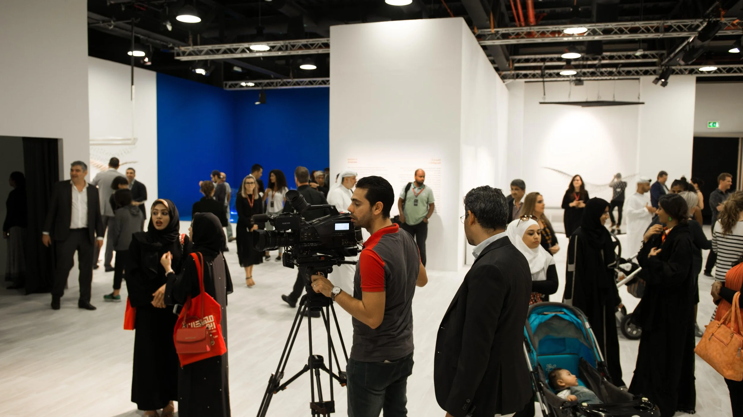 A group of people at an indoor exhibition or conference, some in business attire, some wearing traditional Middle Eastern clothing, talking and walking around. A cameraman records the event, and a young child is in a stroller.