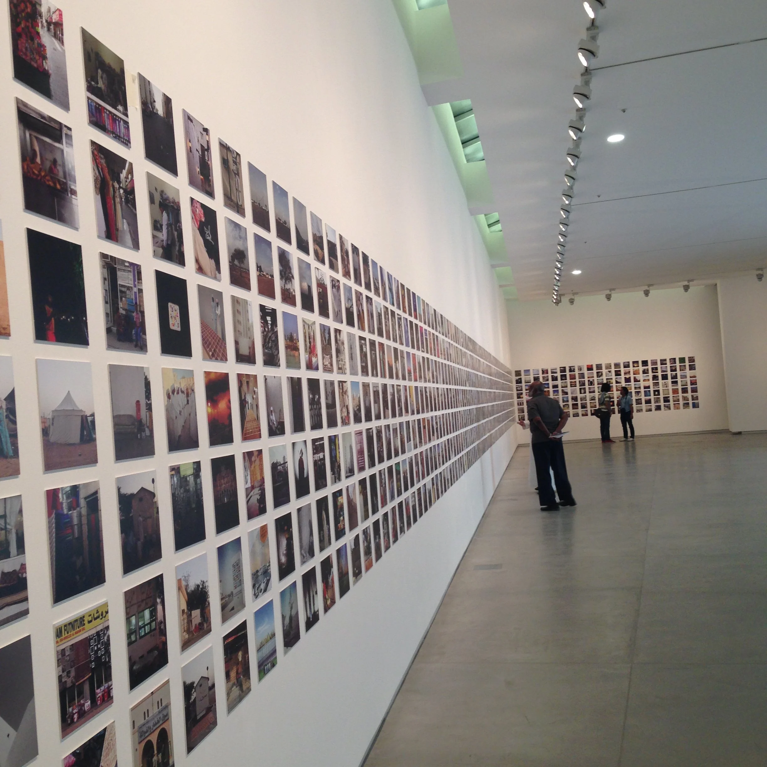 Gallery wall covered with numerous small photographs in an art exhibition, with a few visitors observing.