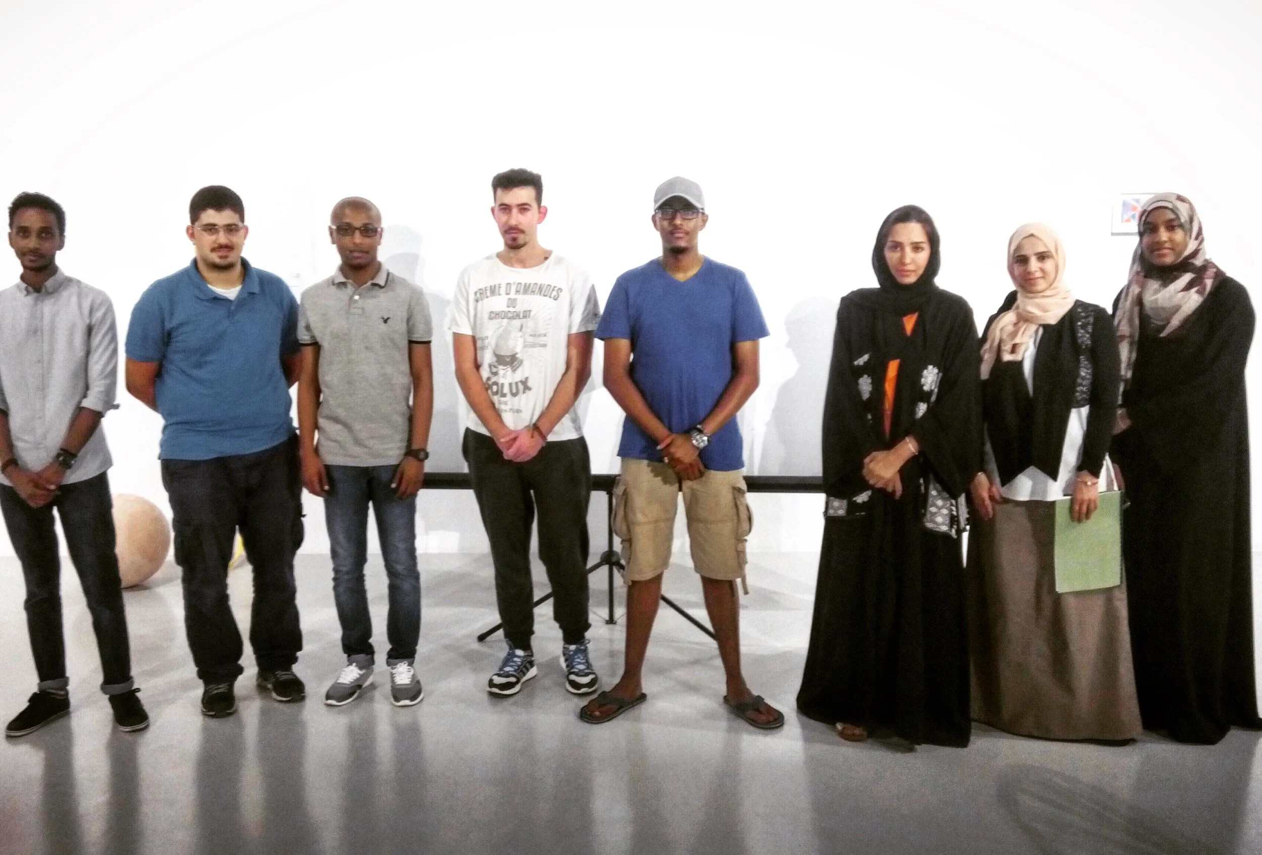 Group of nine diverse young adults standing in a line indoors against a plain white wall, some wearing casual clothing and headscarves, others with glasses, with a music stand in the background.