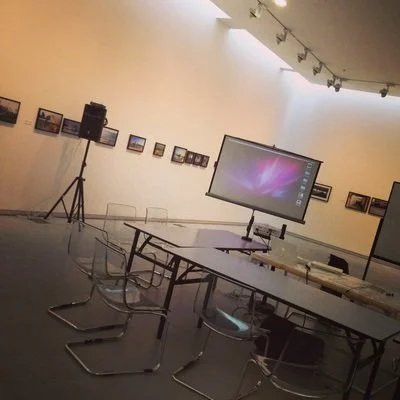 An empty conference room with a large monitor on a table, chairs around the table, and artwork on the walls in an art gallery setting.