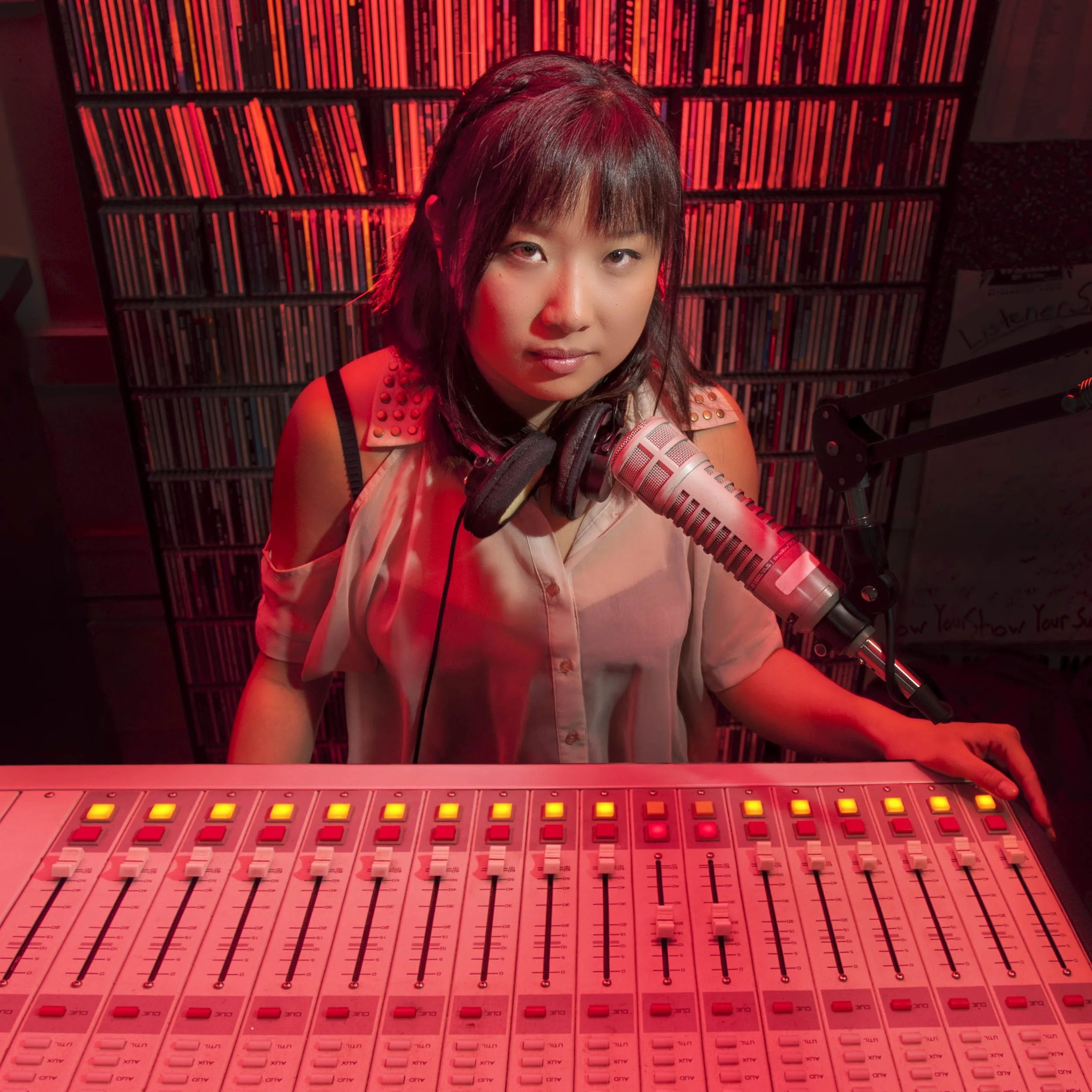 A woman in a recording studio sitting at a sound mixing board with a microphone and over-ear headphones around her neck, with a background of shelves filled with CDs, illuminated by red lighting.